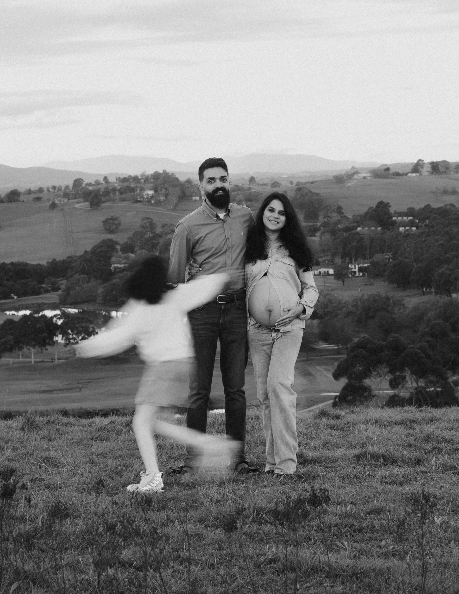 Black and white maternity photo of couple posing on hillside as blurred child runs toward them at dusk.