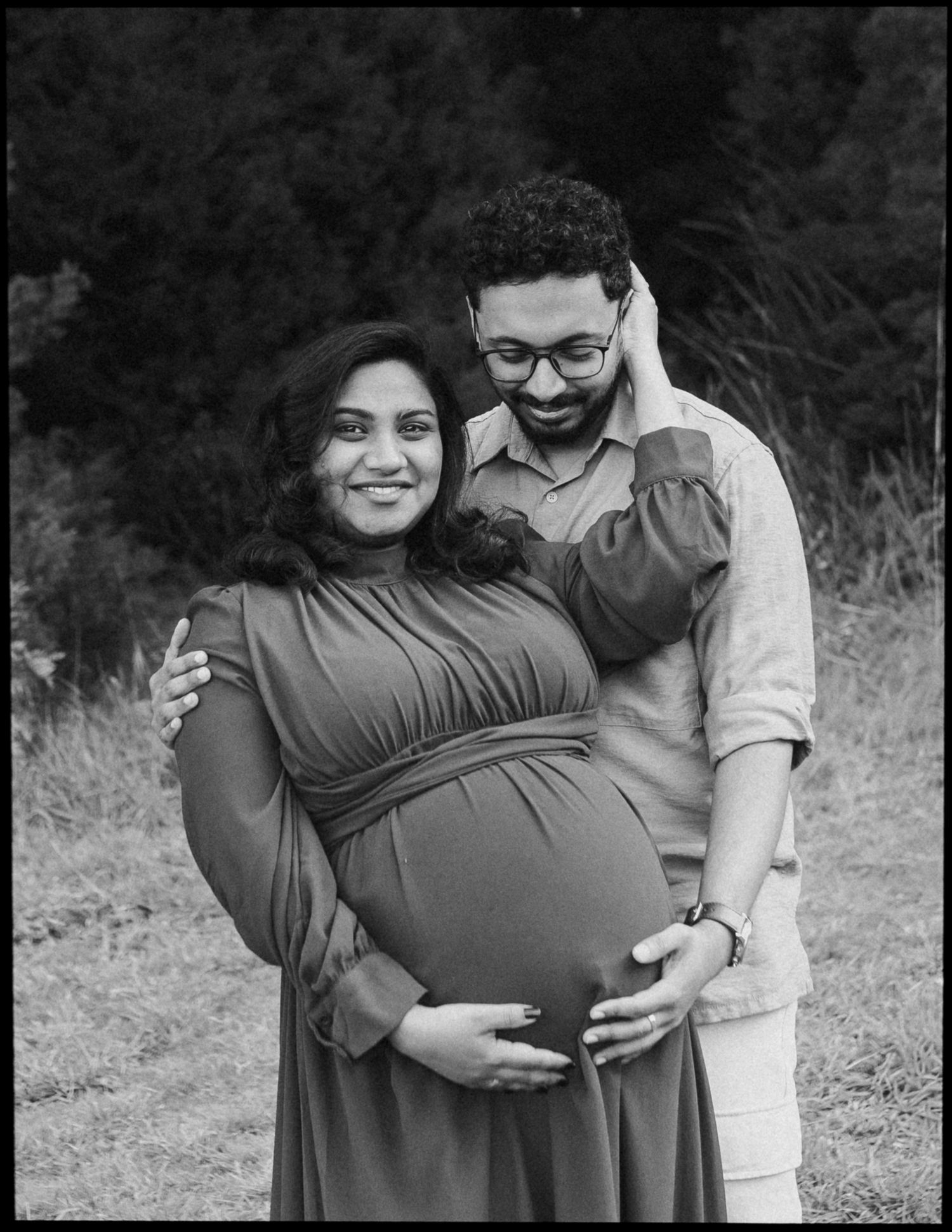 Black and white maternity portrait of a smiling pregnant woman in a long dress embraced from behind by her bearded partner outdoors.
