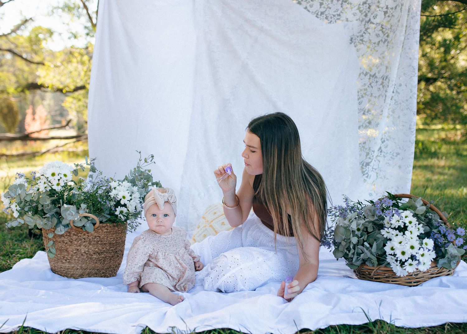 Mother and baby girl sitting together on white blanket surrounded by white flowers.