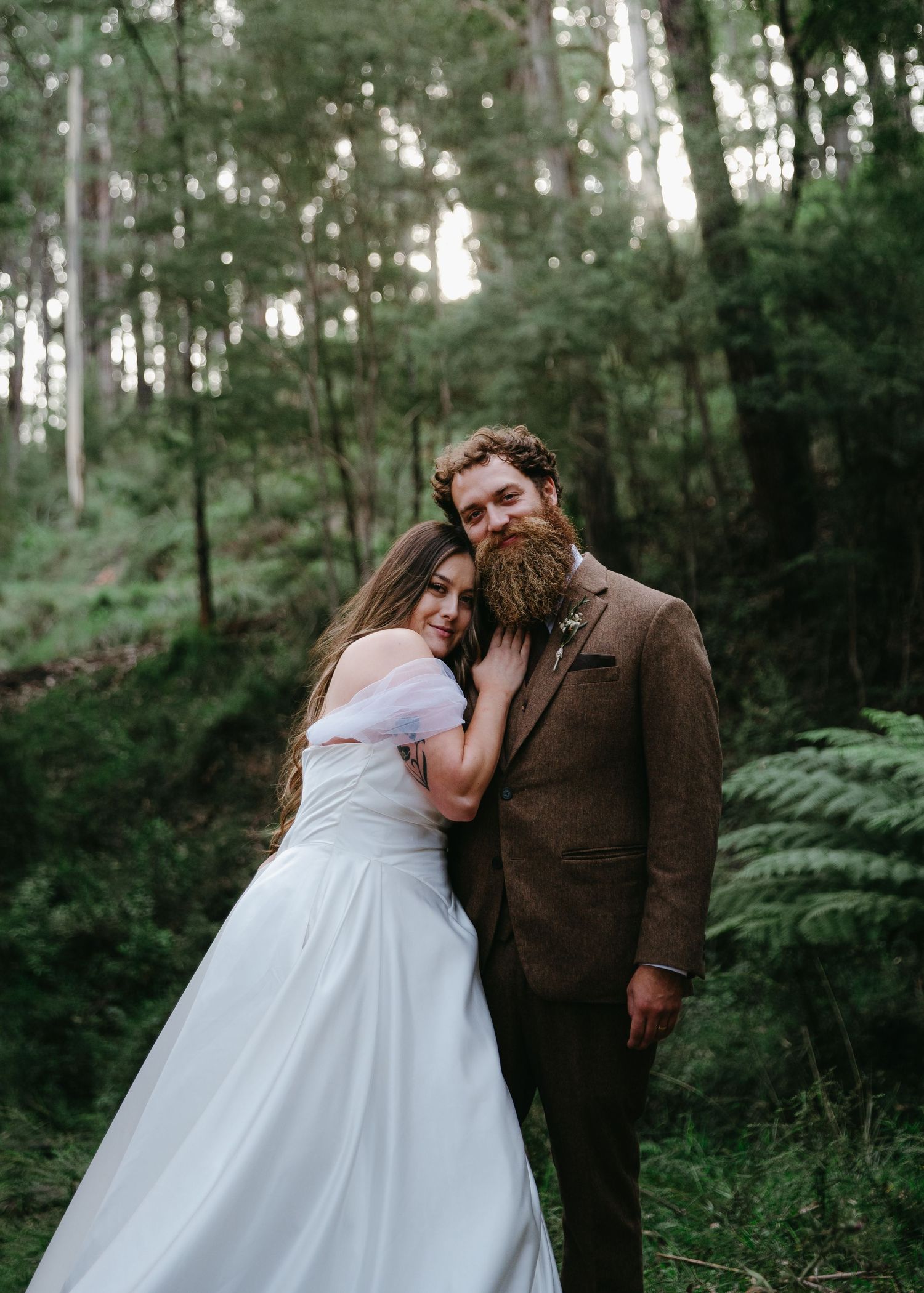 Bride leans on bearded groom shoulder posing in lush green forest.