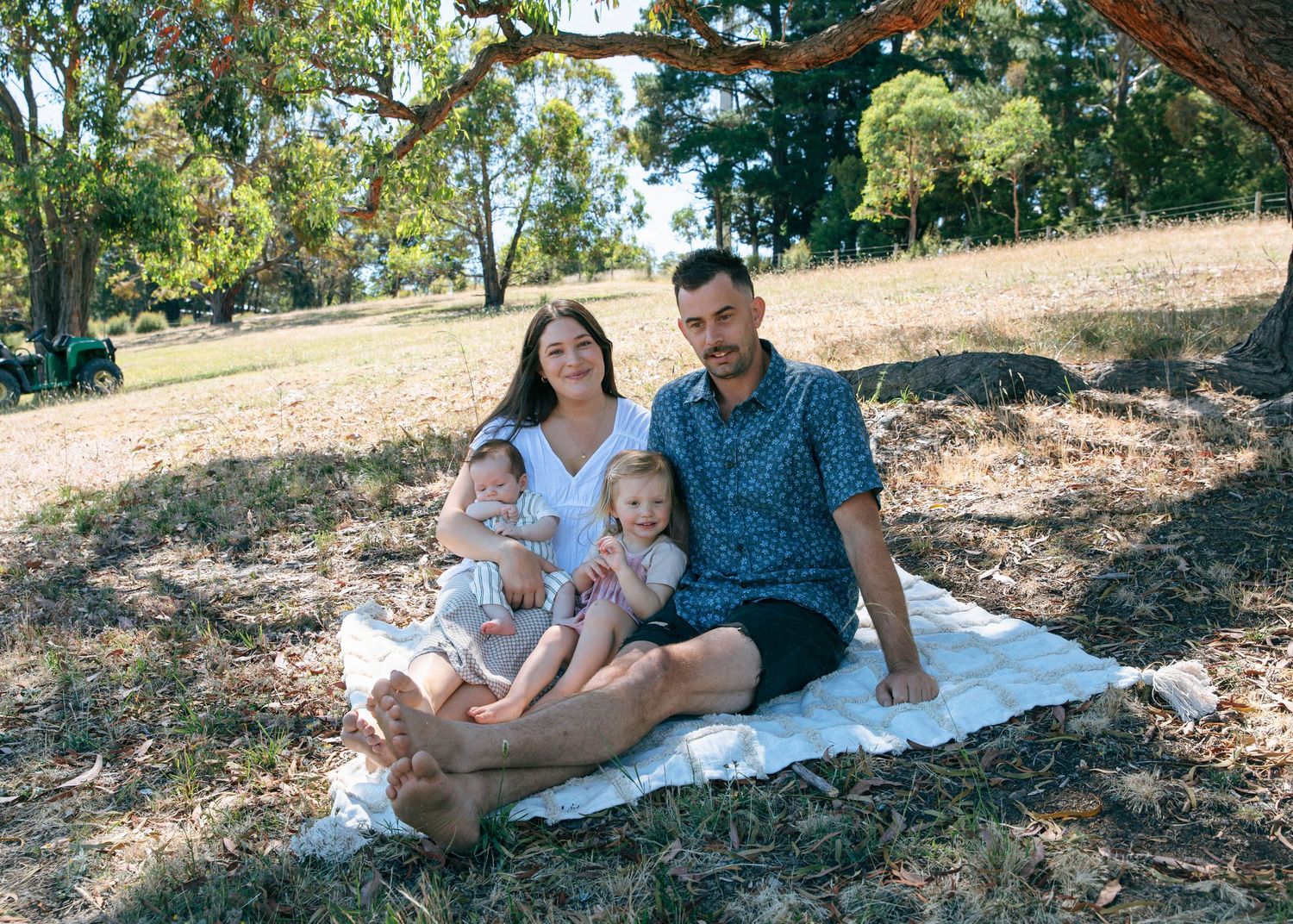 A family of four sits together on a white blanket on dry grass under a tree in a park.