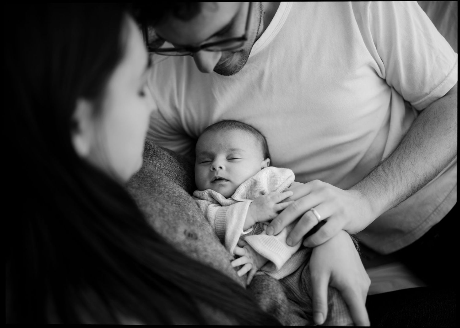 Black and white photo of parents tenderly cradling their sleeping newborn baby together.