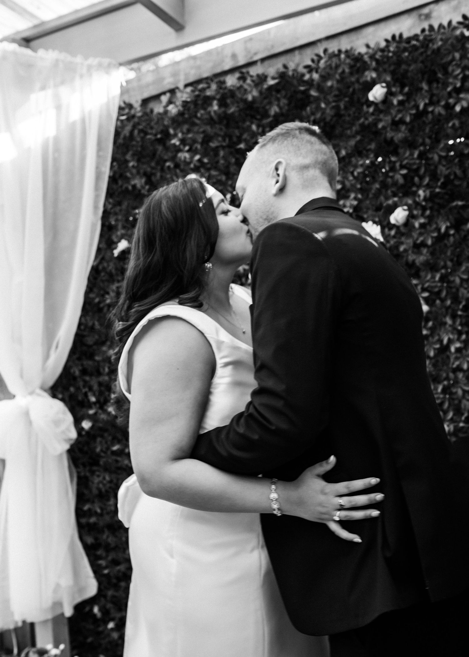 Black and white photo of newlyweds sharing first kiss at outdoor wedding ceremony with floral backdrop.