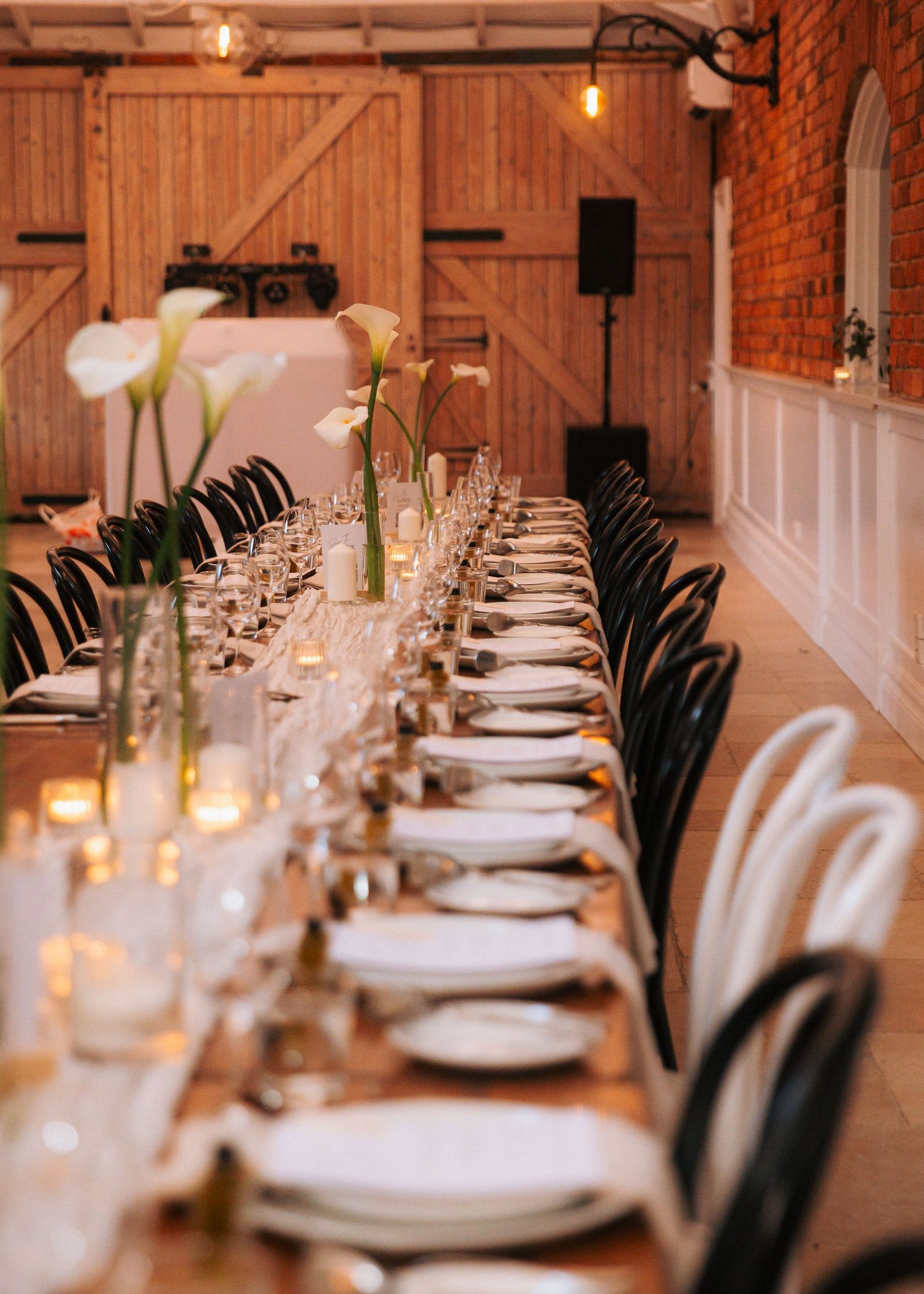 Elegant barn wedding reception with long wooden table, white floral centerpieces, candles, and mixed black and white chairs.