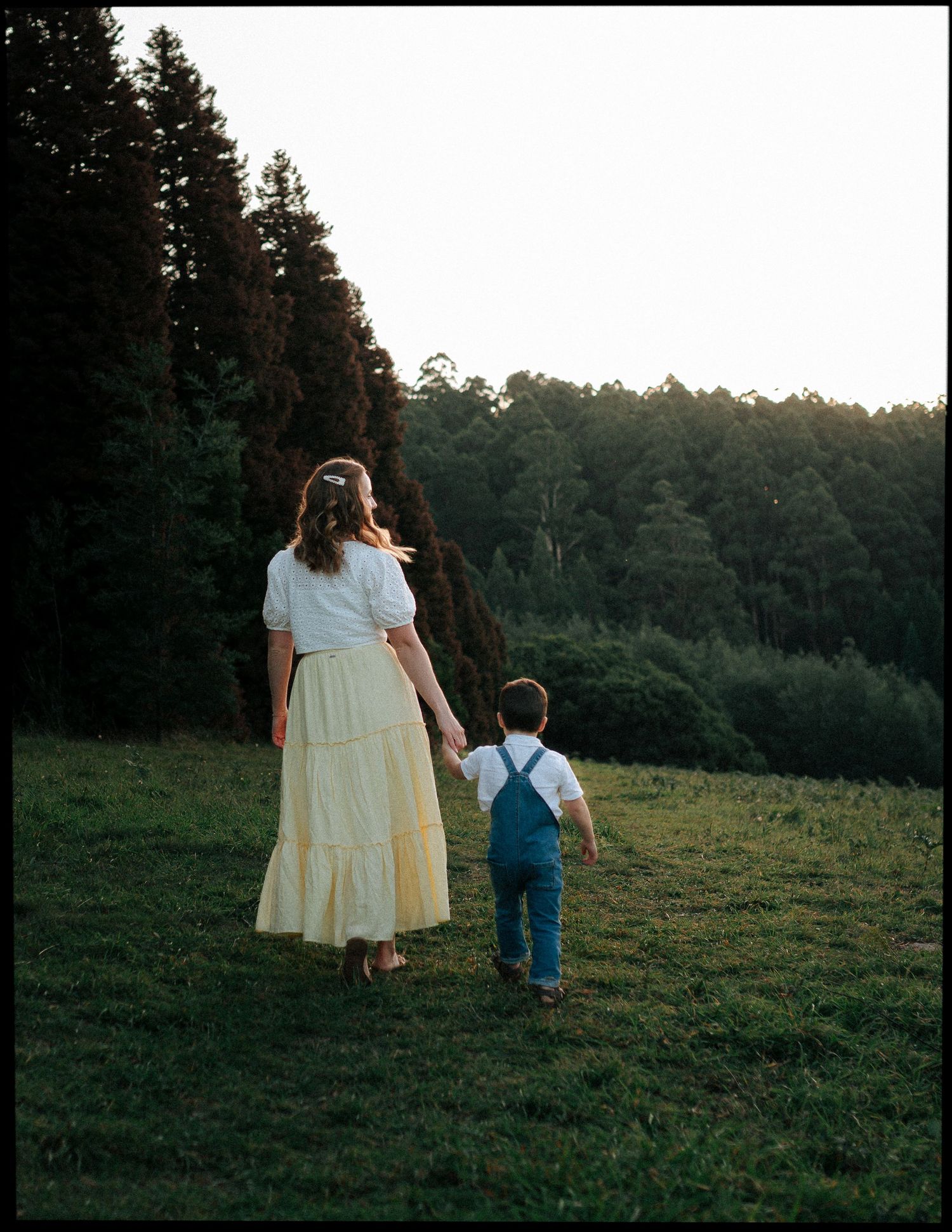 Mother and toddler son holding hands walking toward tall evergreen forest in golden evening light.