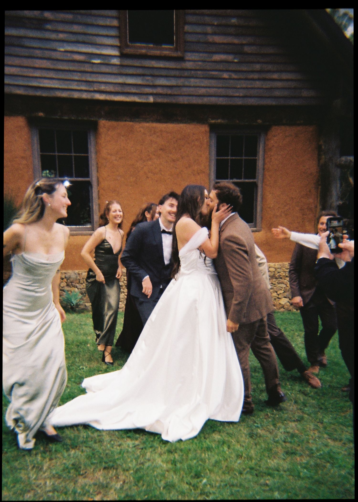 Bride and groom share a kiss outdoors surrounded by smiling wedding guests near rustic building.