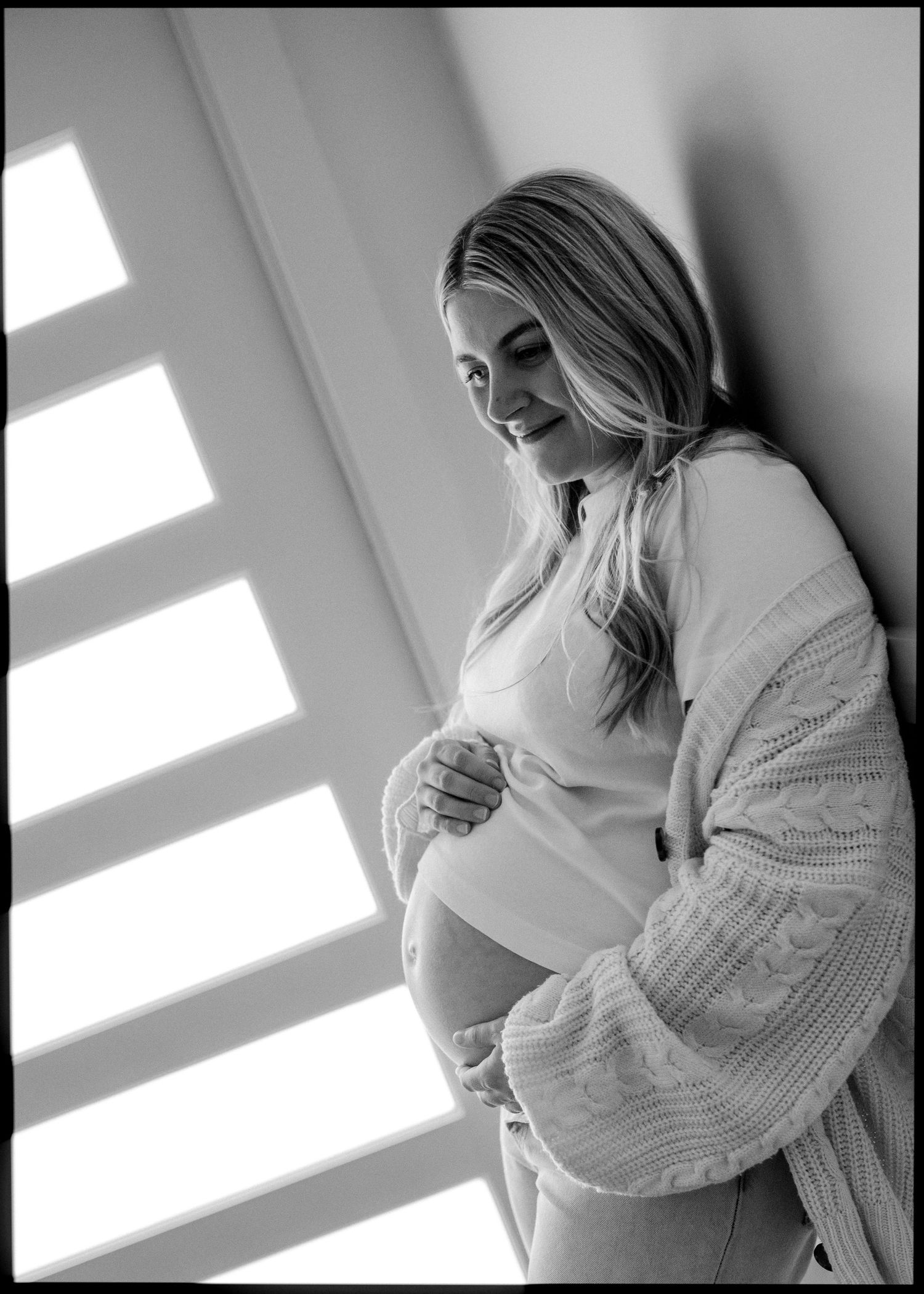 Black and white portrait of smiling pregnant woman cradling bump in cozy cardigan by shuttered window.