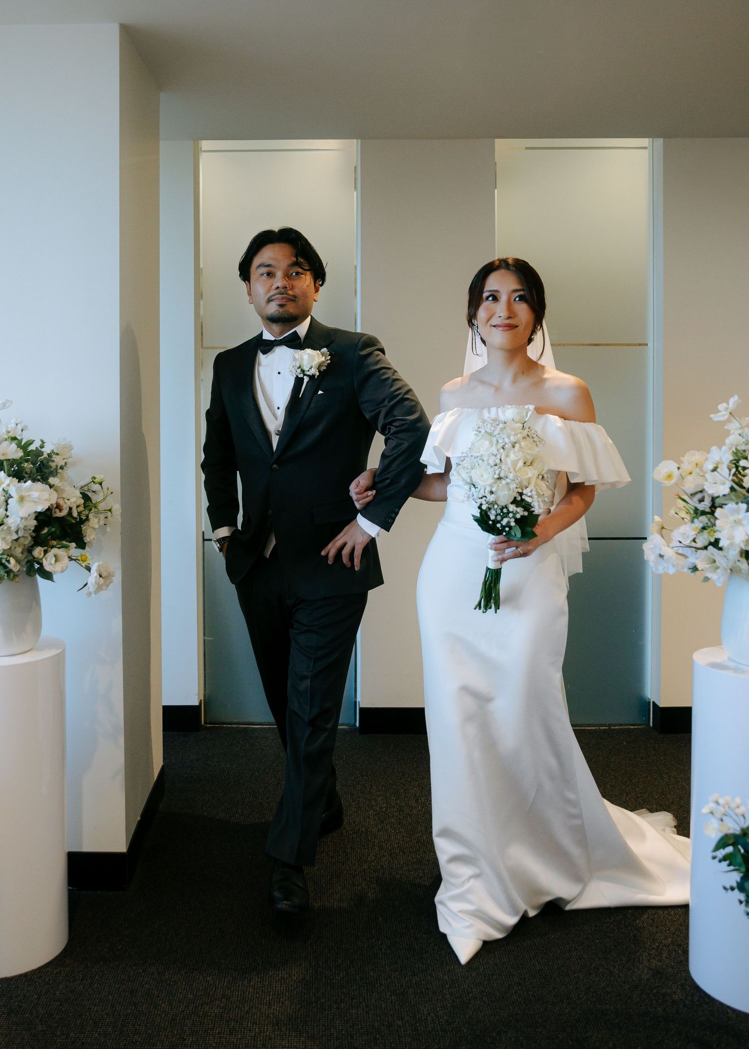 Bride in off-shoulder white gown and groom in black tuxedo walking together at wedding ceremony.
