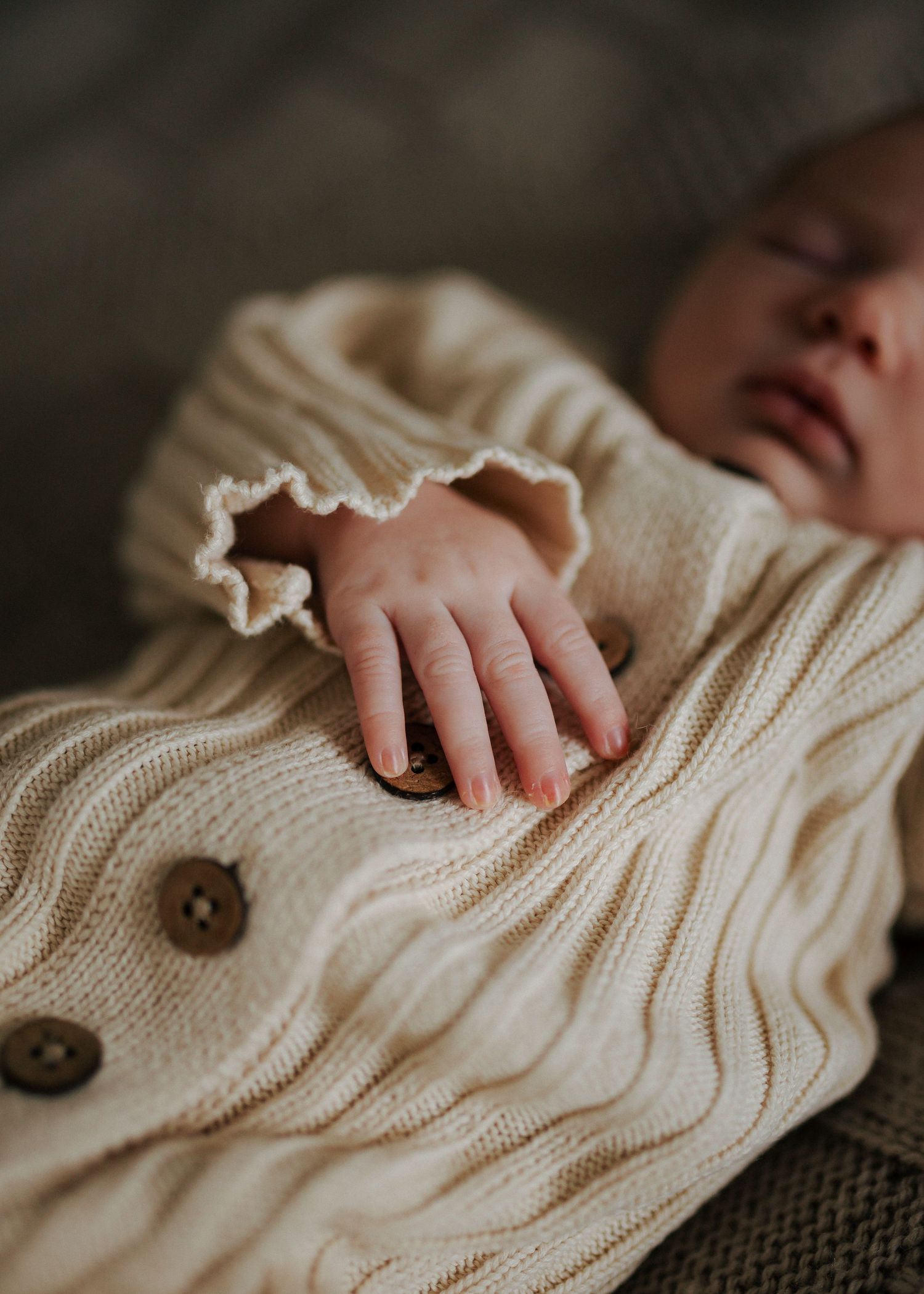 Macro shot of newborn baby tiny hand resting on cream cable-knit button cardigan while sleeping.