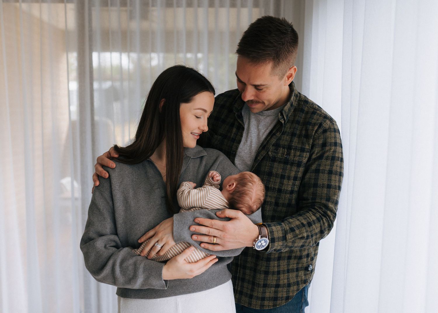 New parents lovingly cradle their sleeping newborn baby together near a bright window.