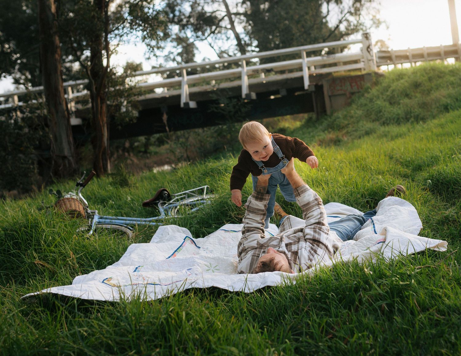Father lying on a blanket in green grass playing with happy toddler near a white bridge outdoors.