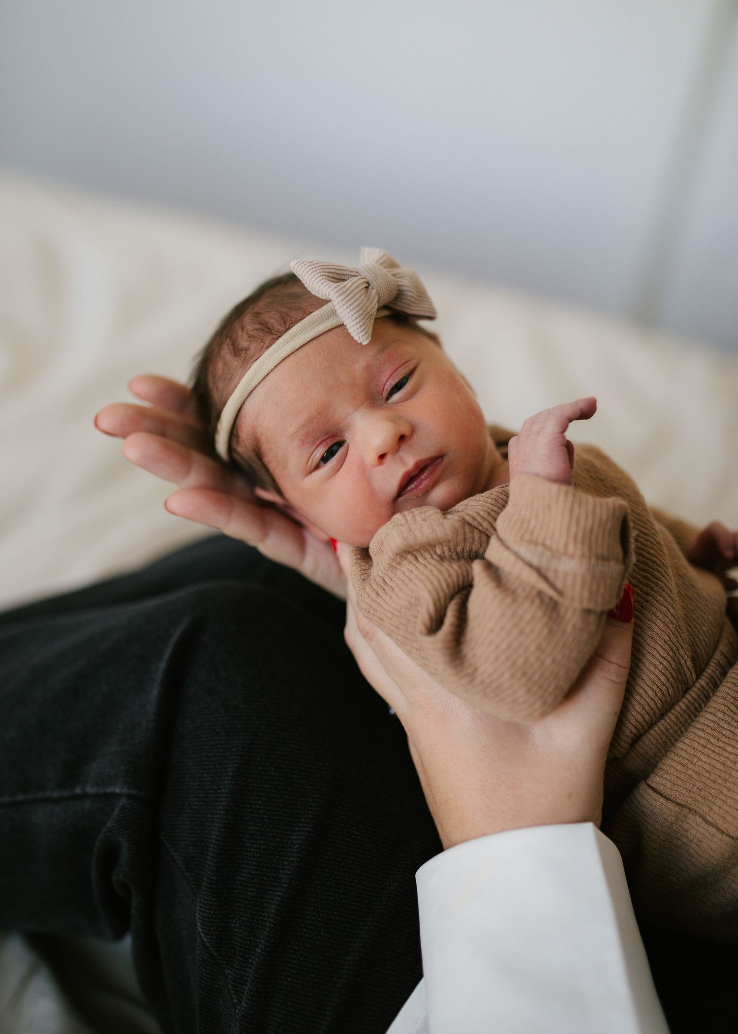 Newborn baby girl in brown romper with beige bow headband cradled in parent's hands.