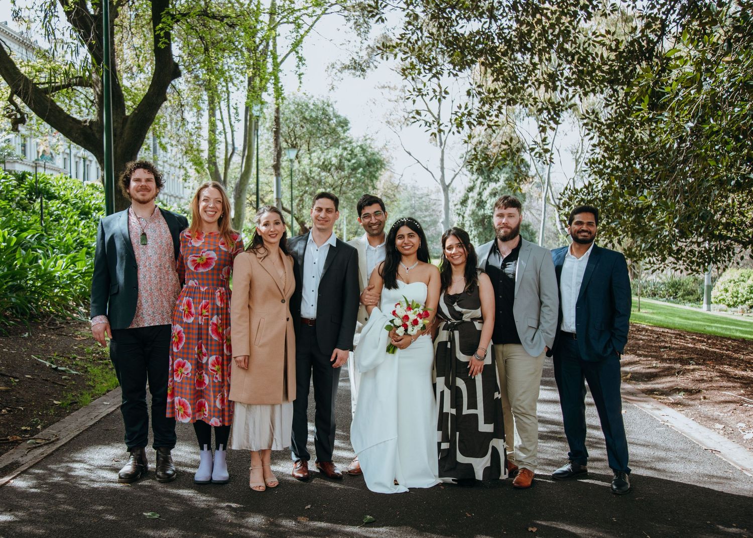 Wedding party group photo with bride in white gown and groom posing with friends in a lush green park setting.