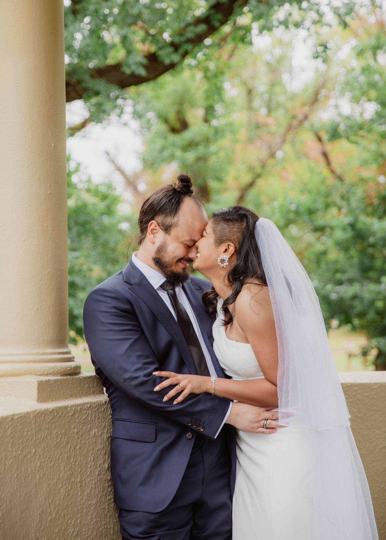 Bride and groom sharing an intimate moment between stone columns in a park.