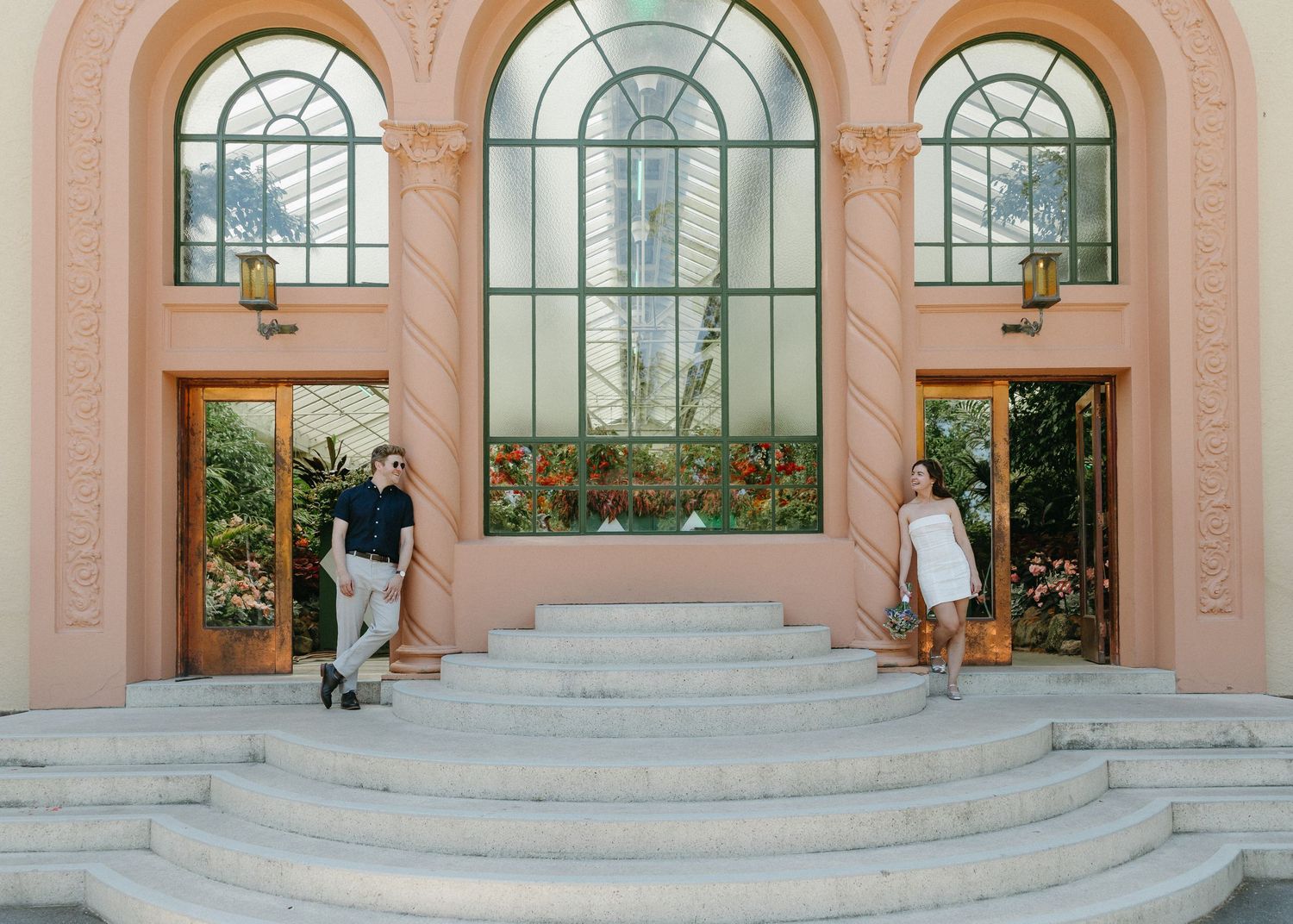 Bride and groom descend marble steps of elegant pink building with arched glass windows.
