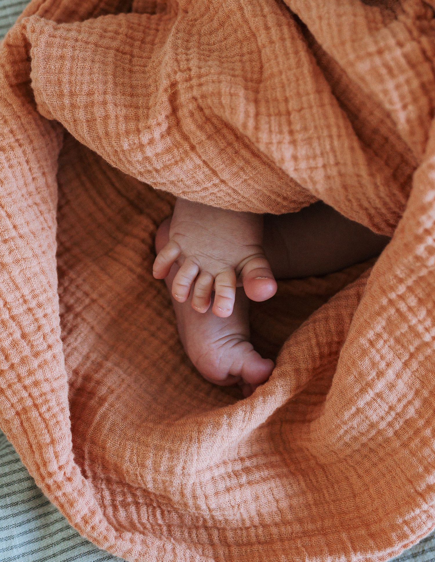 A newborn baby's tiny bare feet peek out from a soft, textured peach muslin blanket resting on a striped surface.