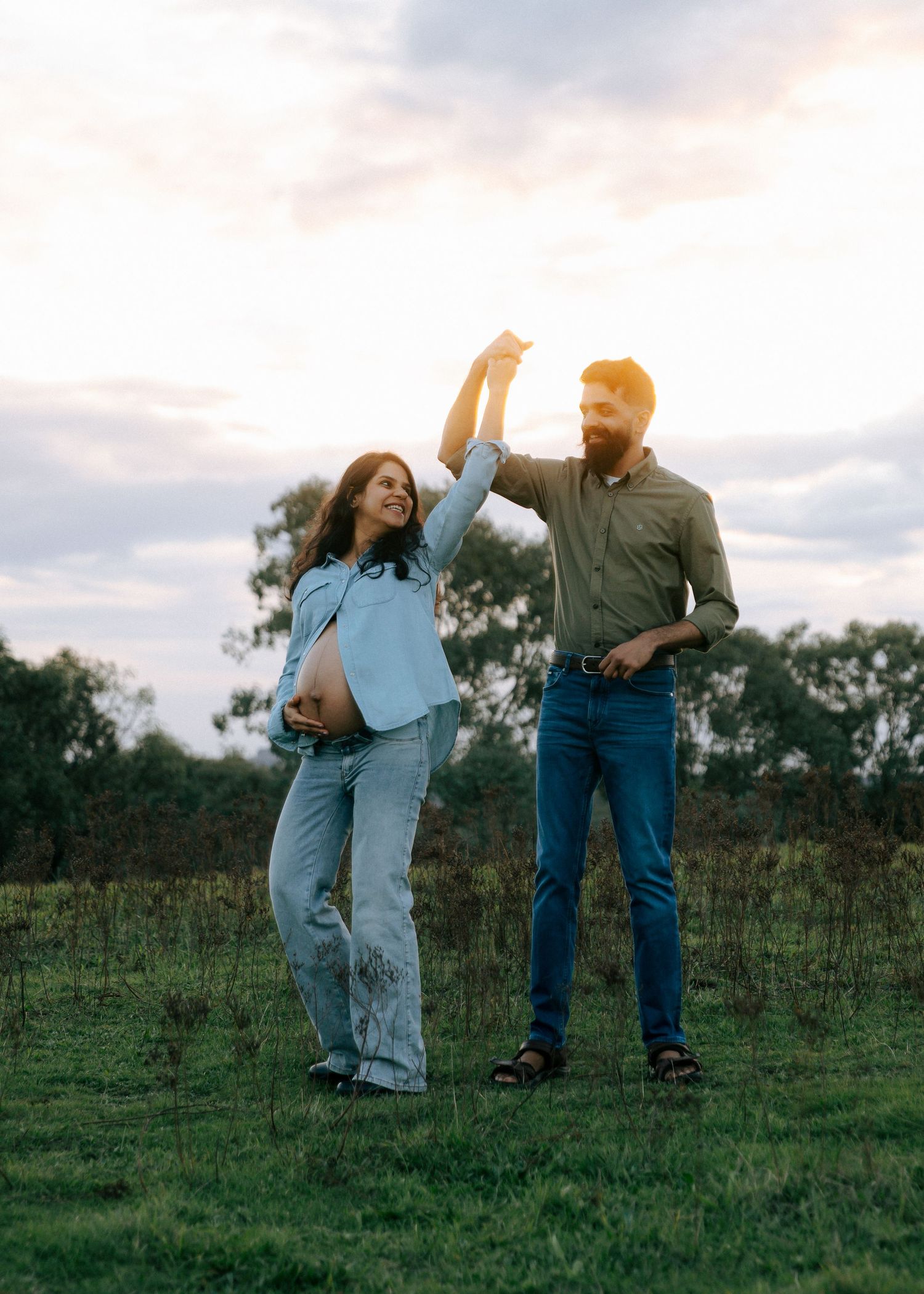 Pregnant couple dancing joyfully on a grassy field at golden hour with trees in background.