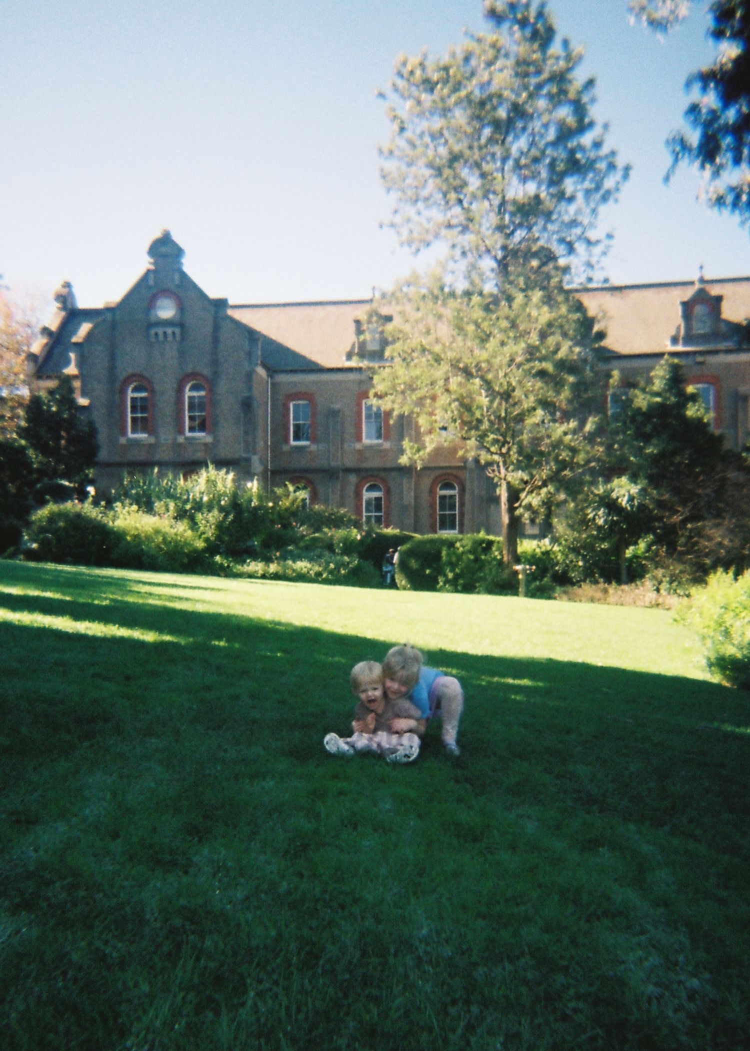 Two toddlers play together on a lush green lawn in front of a grand historic stone building.