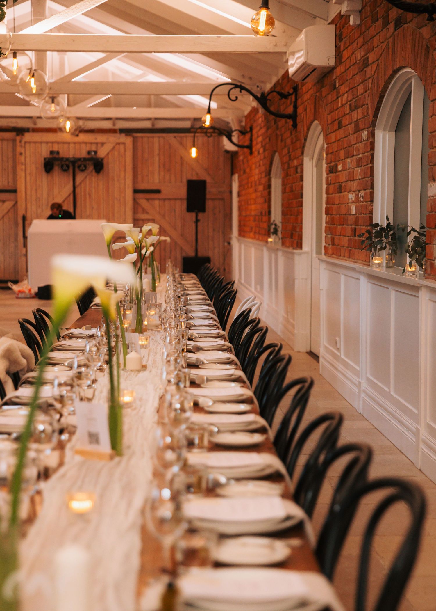 Elegant wedding reception long table set with white plates, glassware, and calla lily centerpieces in brick venue.