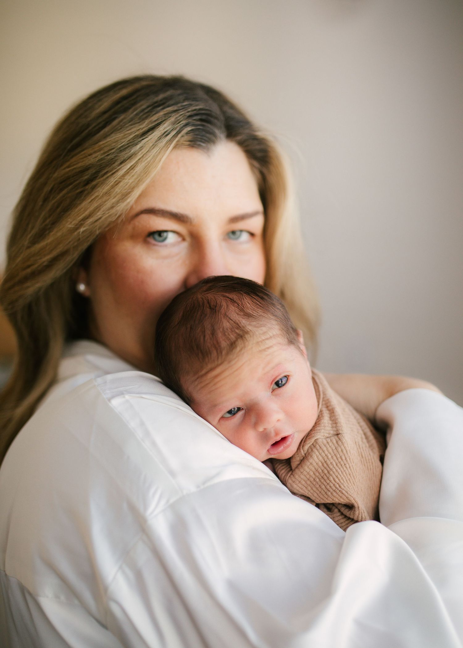 Mother in white shirt tenderly holds newborn baby close to her chest in a soft natural light portrait.