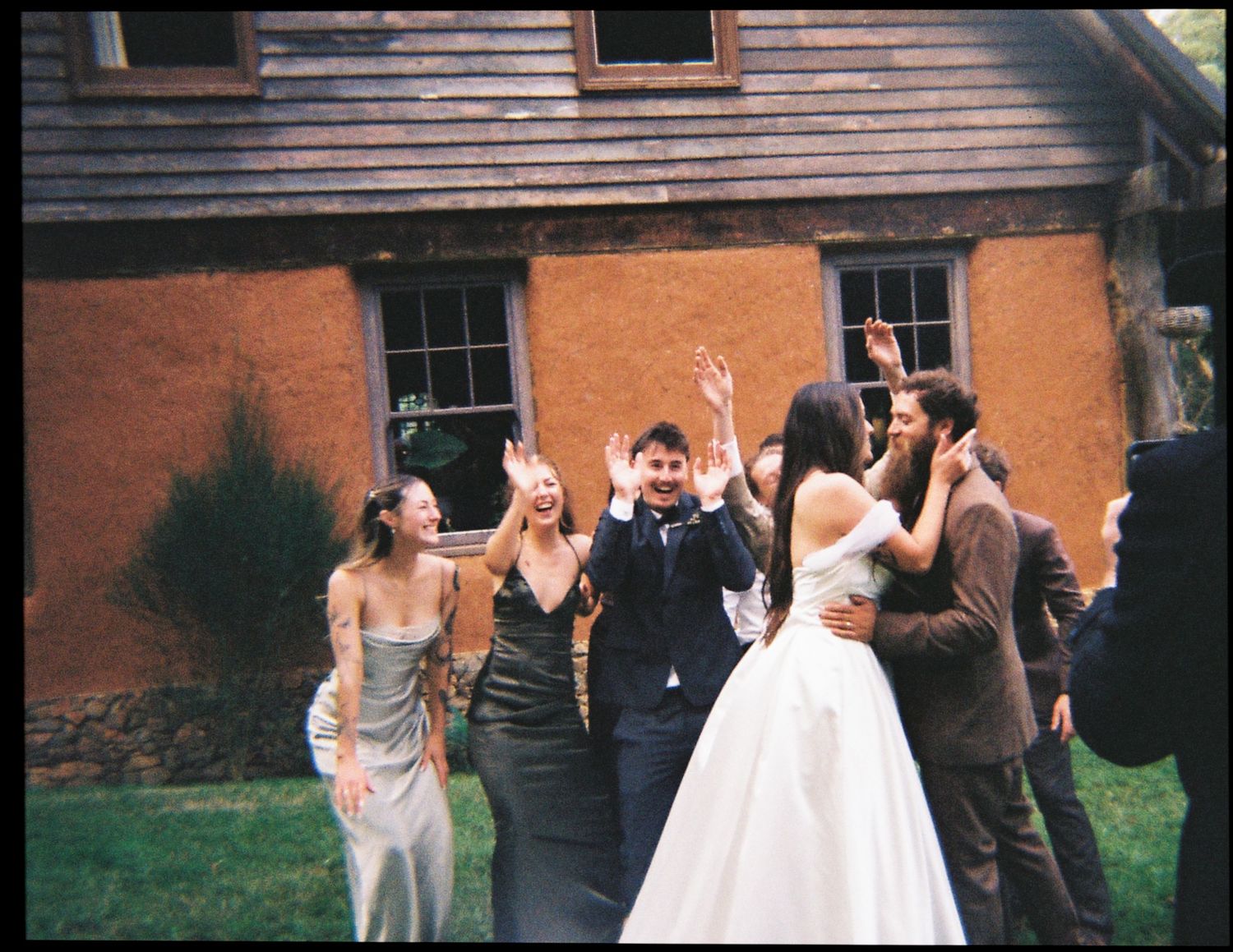 Joyful wedding party celebrating outside a rustic brick building, with guests cheering and embracing the happy couple.