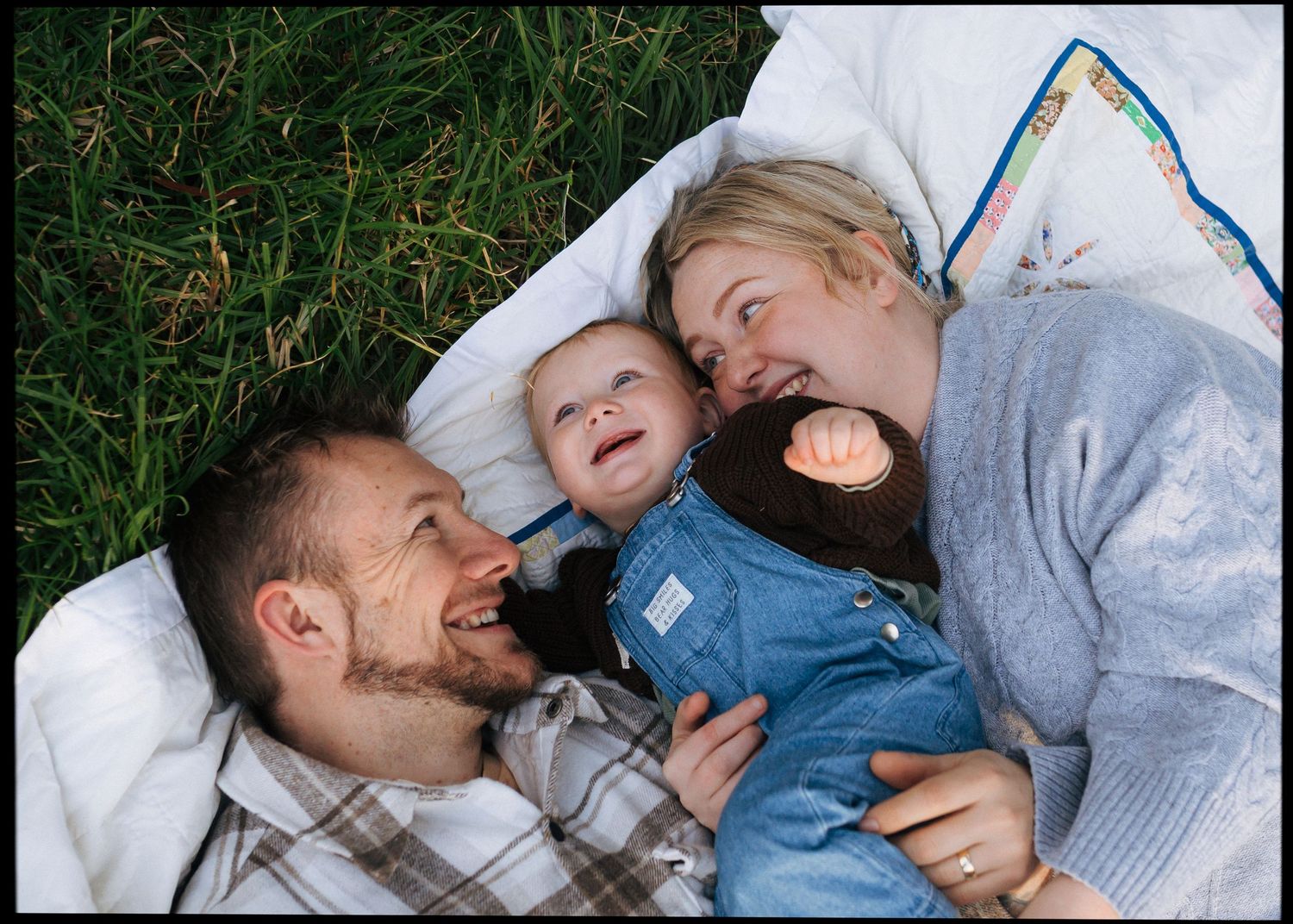 Happy family of three lying on a blanket in the grass, smiling and laughing together outdoors.