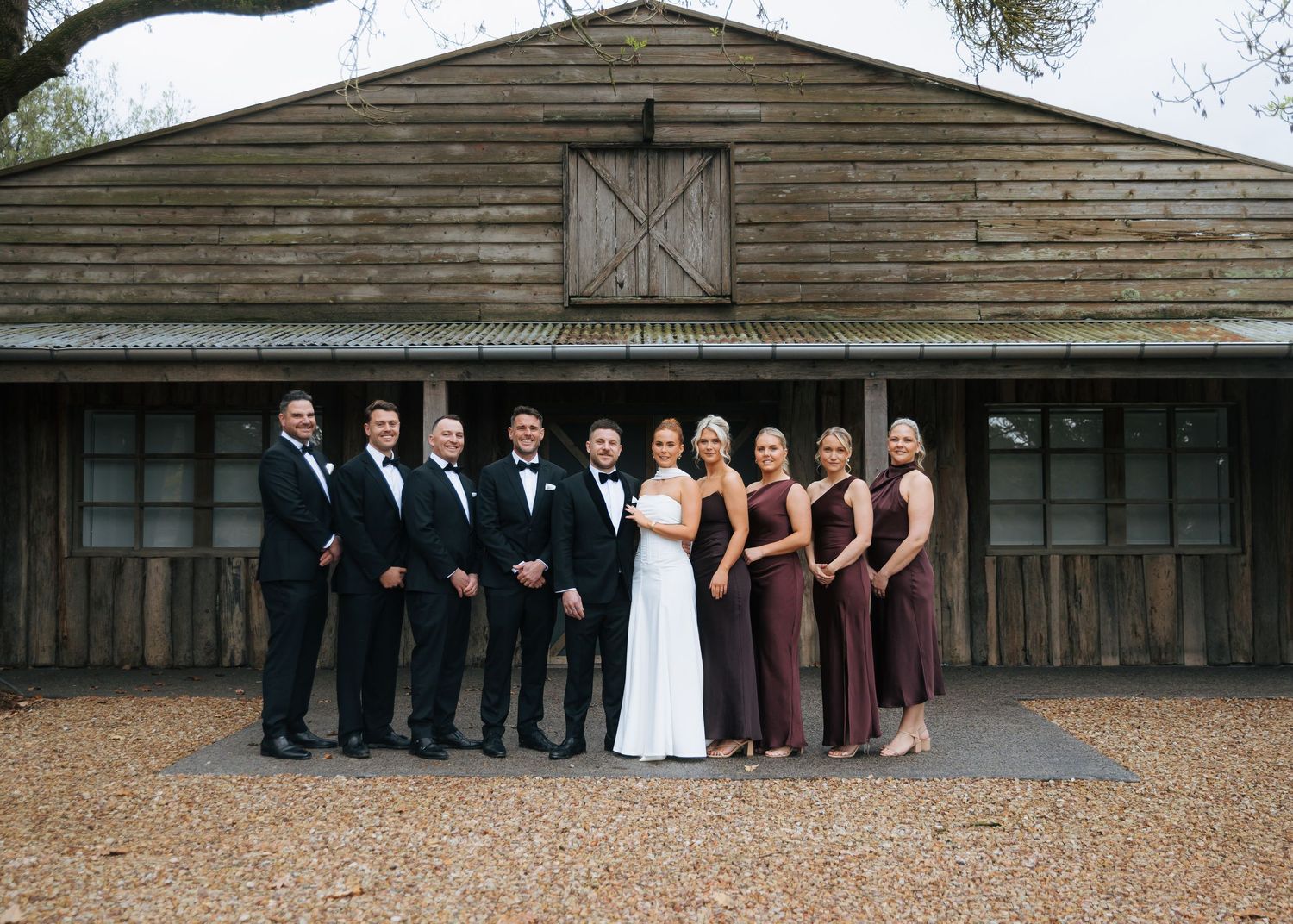 Wedding party in black tuxedos and burgundy gowns pose before rustic wooden barn.