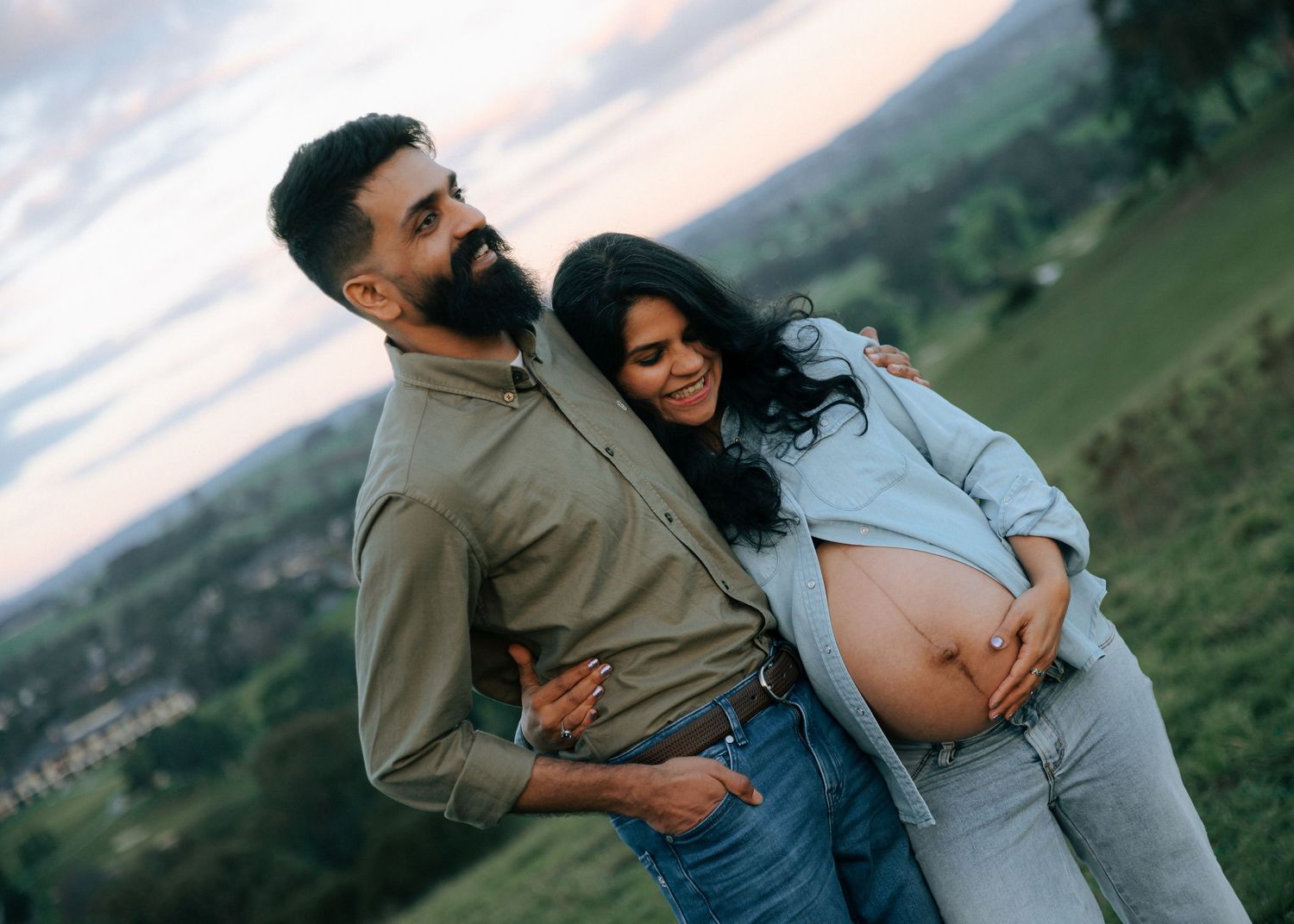 Happy expecting couple snuggling close at sunset with lush green countryside landscape behind them.