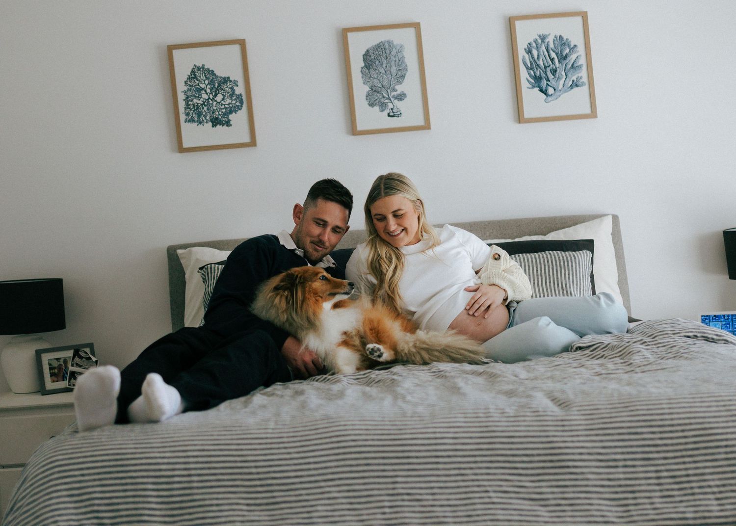 Couple relaxing on bed with fluffy dog, pregnant woman in white outfit smiling warmly.