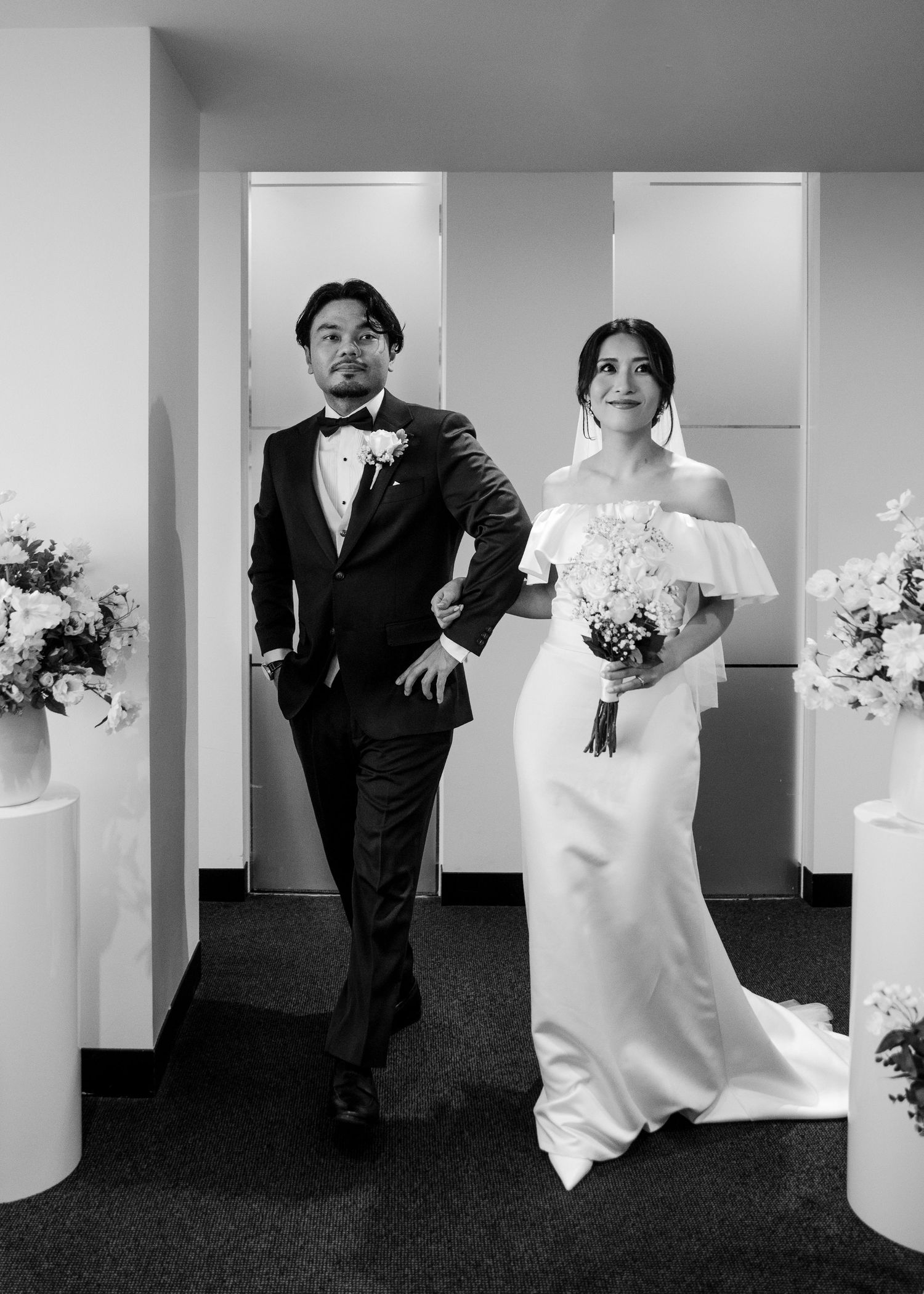 Black and white photo of bride and groom walking together flanked by white floral arrangements.