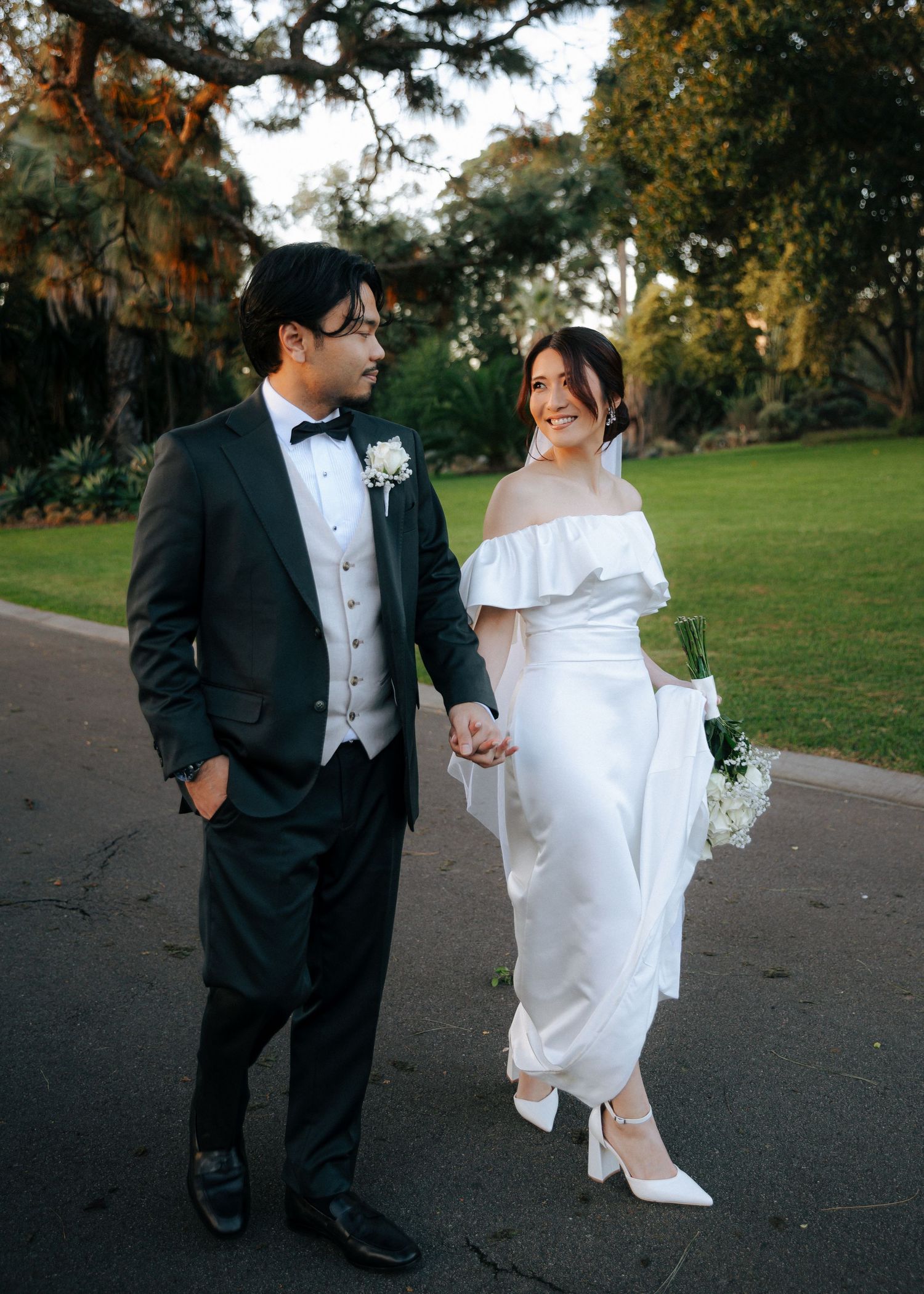 Newlywed couple walking hand in hand outdoors, groom in black tuxedo, bride in white off-shoulder dress.