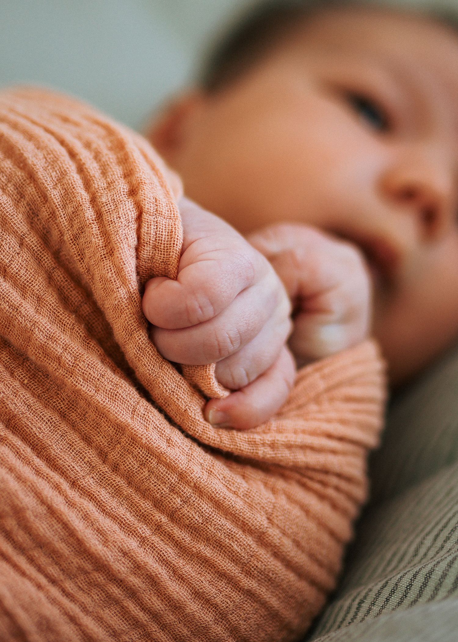 Newborn baby wrapped in a soft orange muslin swaddle with a tiny hand gripping the fabric.