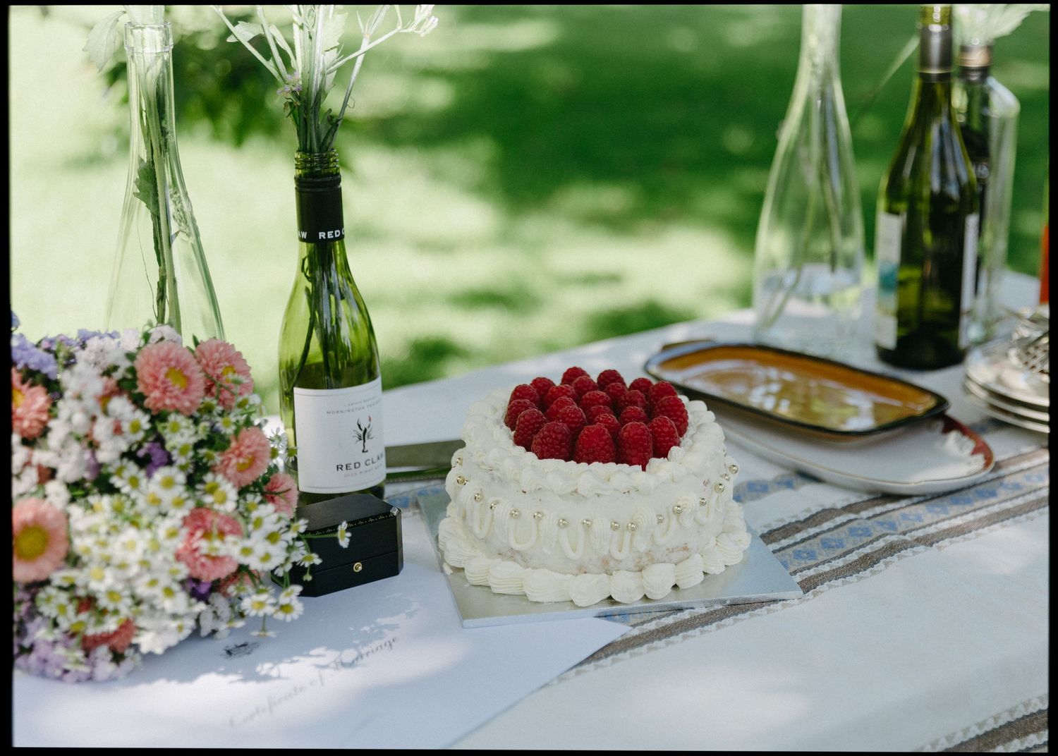 White wedding cake topped with fresh raspberries on an outdoor table with wine bottles and floral arrangements.