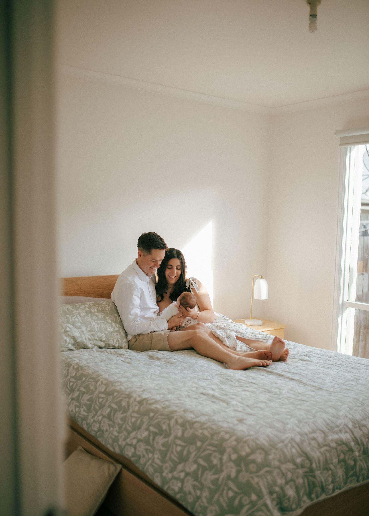 Loving couple sitting on a bed in a sunlit bedroom tenderly holding their newborn baby.