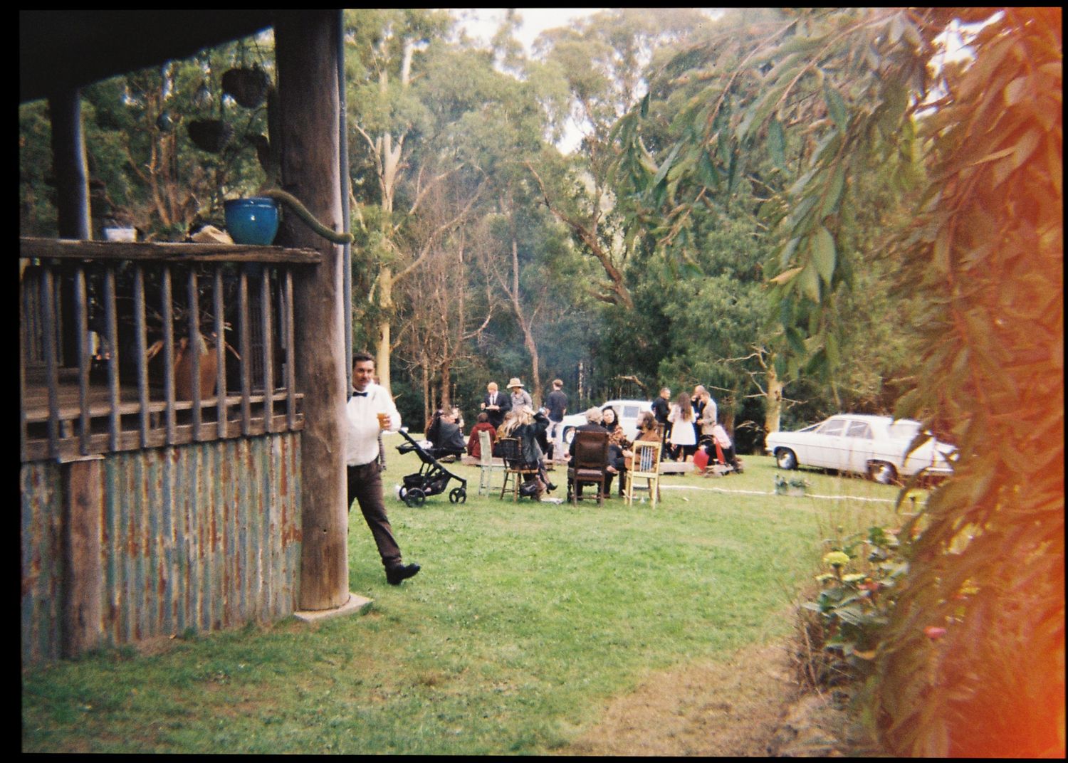 Outdoor wedding reception gathering on a green lawn surrounded by lush trees and natural garden setting.