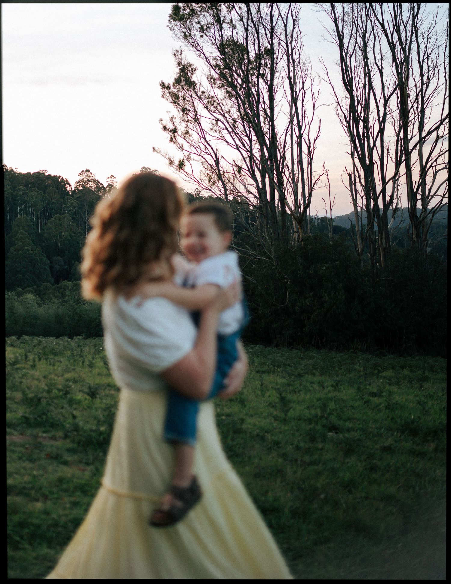 A soft-focus portrait of a mother cradling her toddler son in a lush outdoor evening setting.