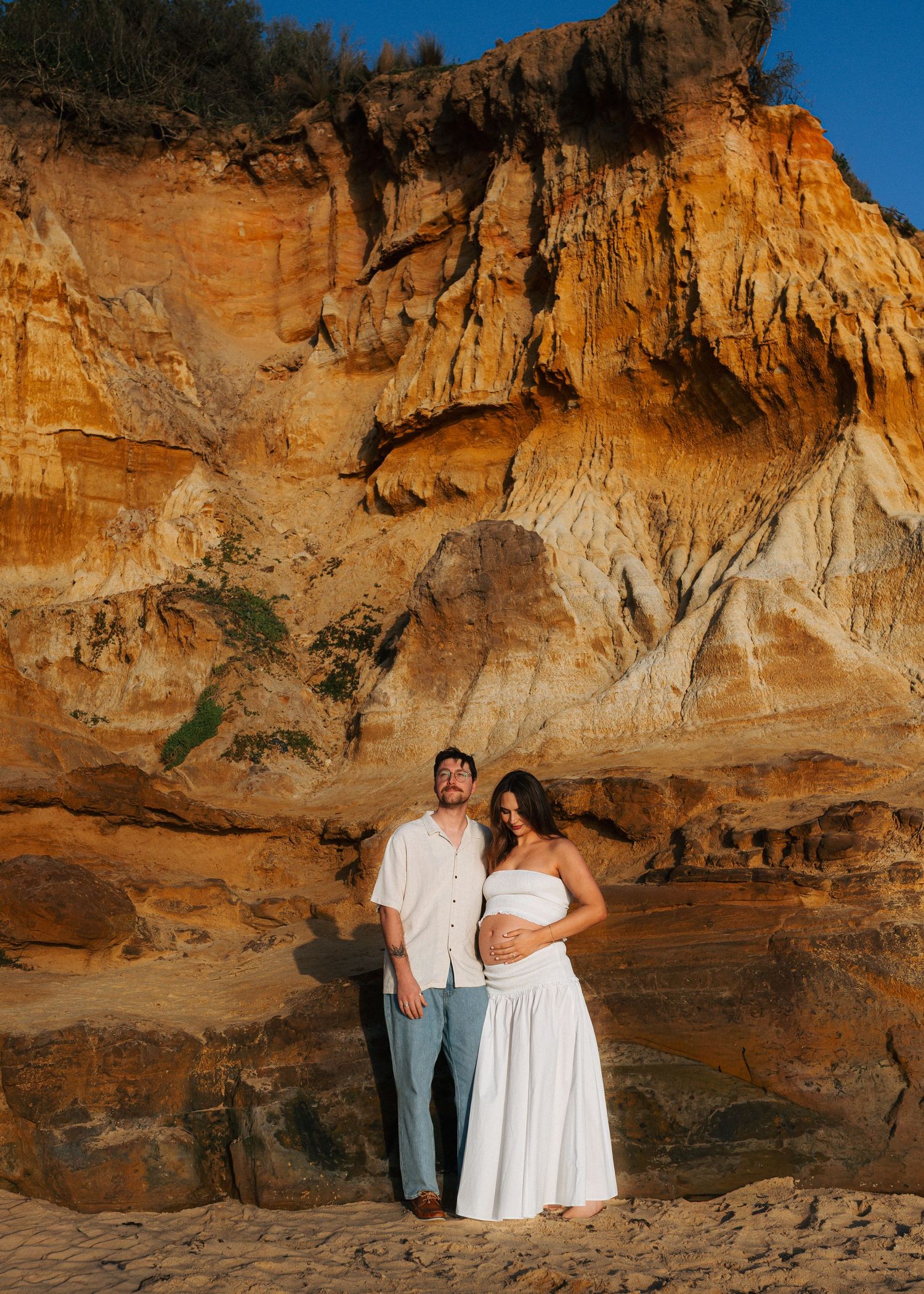 Couple poses before golden sandstone cliffs during maternity shoot, woman in white dress cradling baby bump.