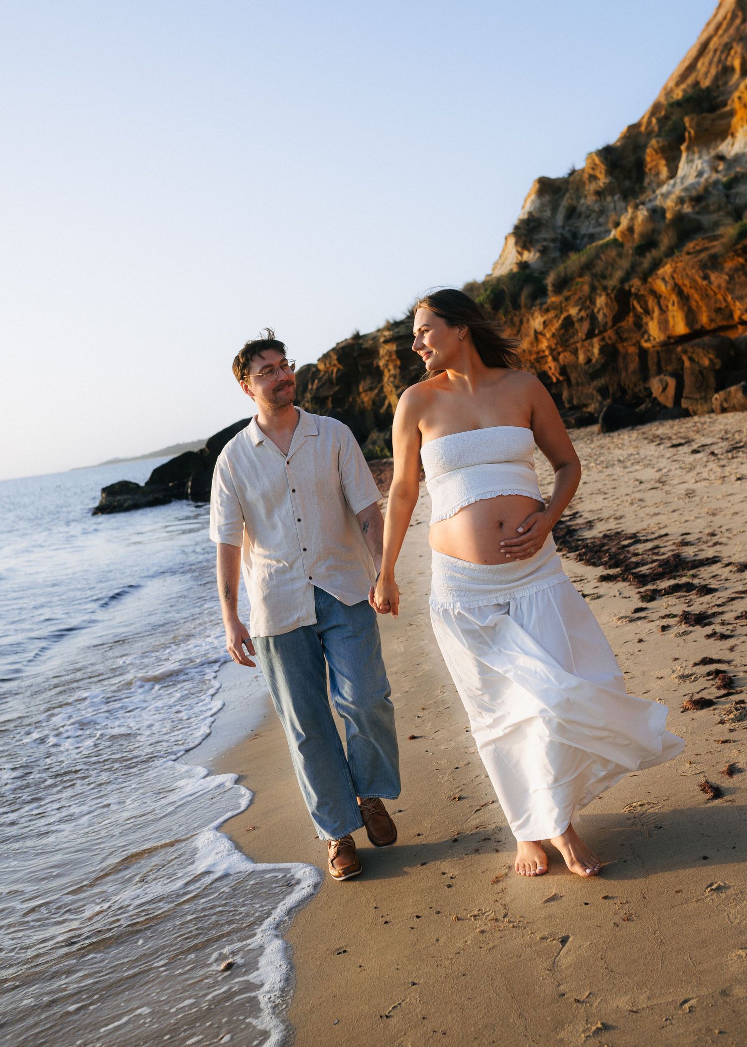 Pregnant couple walking hand-in-hand on a sunny beach, woman in white dress cradling her baby bump near rocky cliffs.