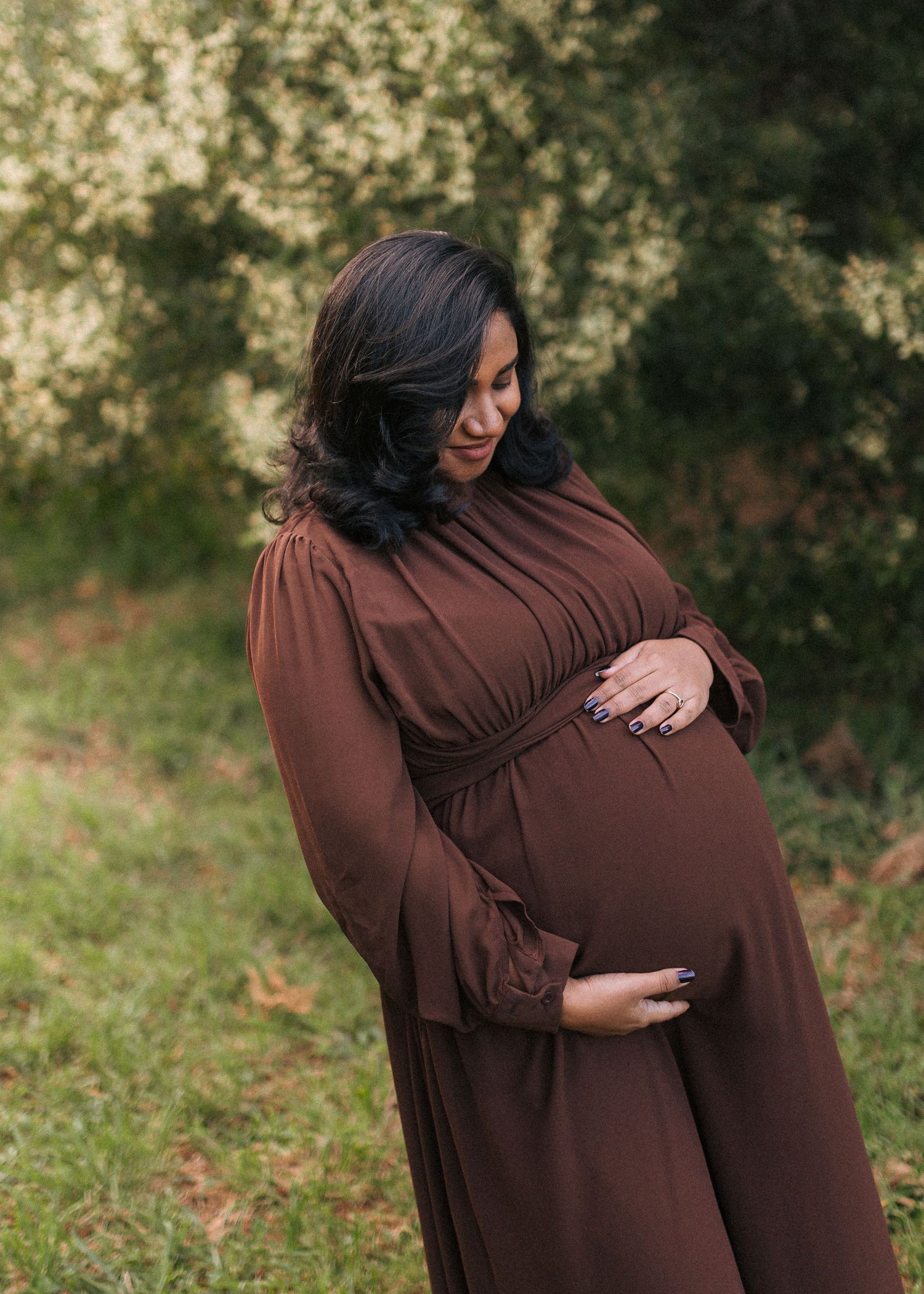 Pregnant woman in a flowing brown dress cradles her baby bump while smiling down in a sunlit green park during a maternity photoshoot.