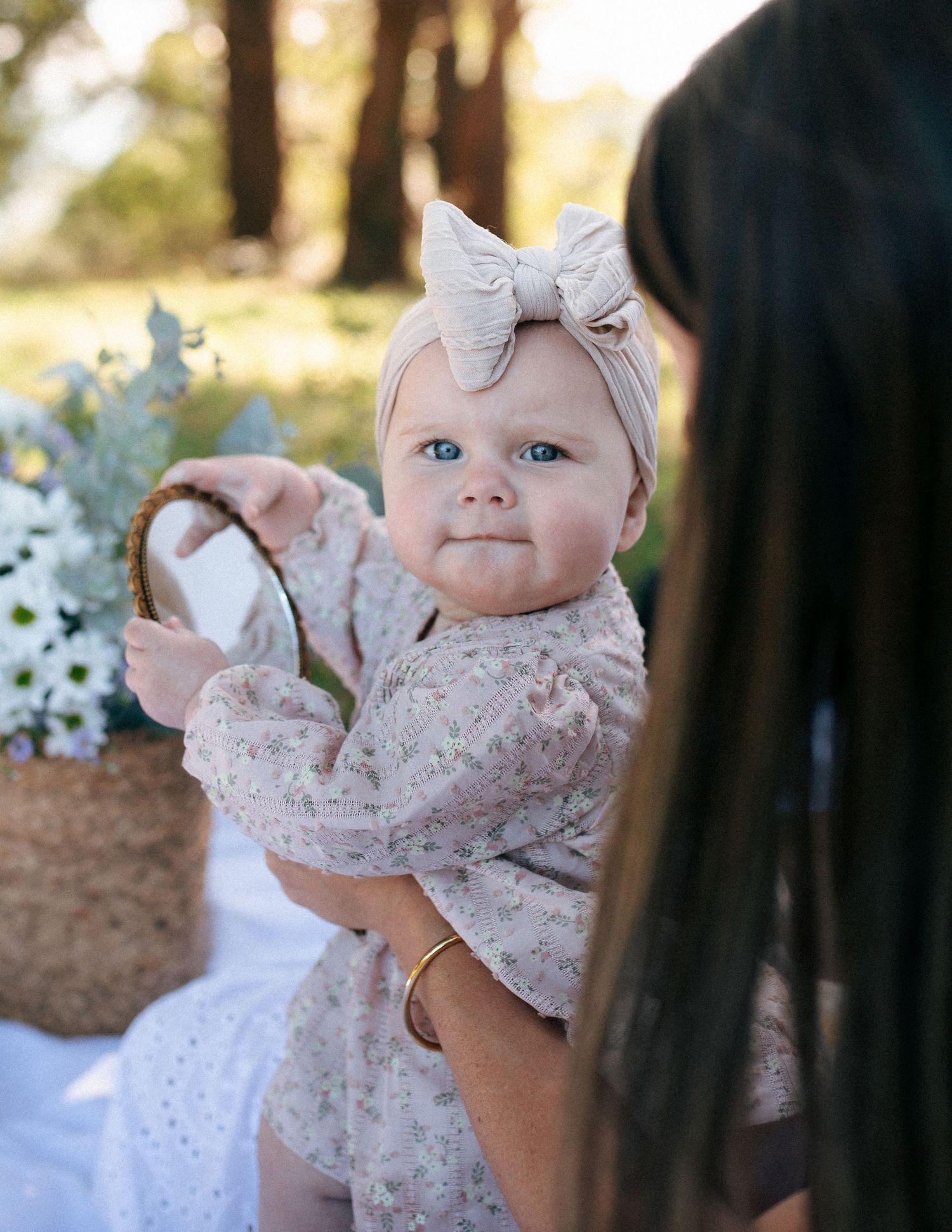 Blue-eyed baby girl in floral dress and bow headband held by mother outdoors.