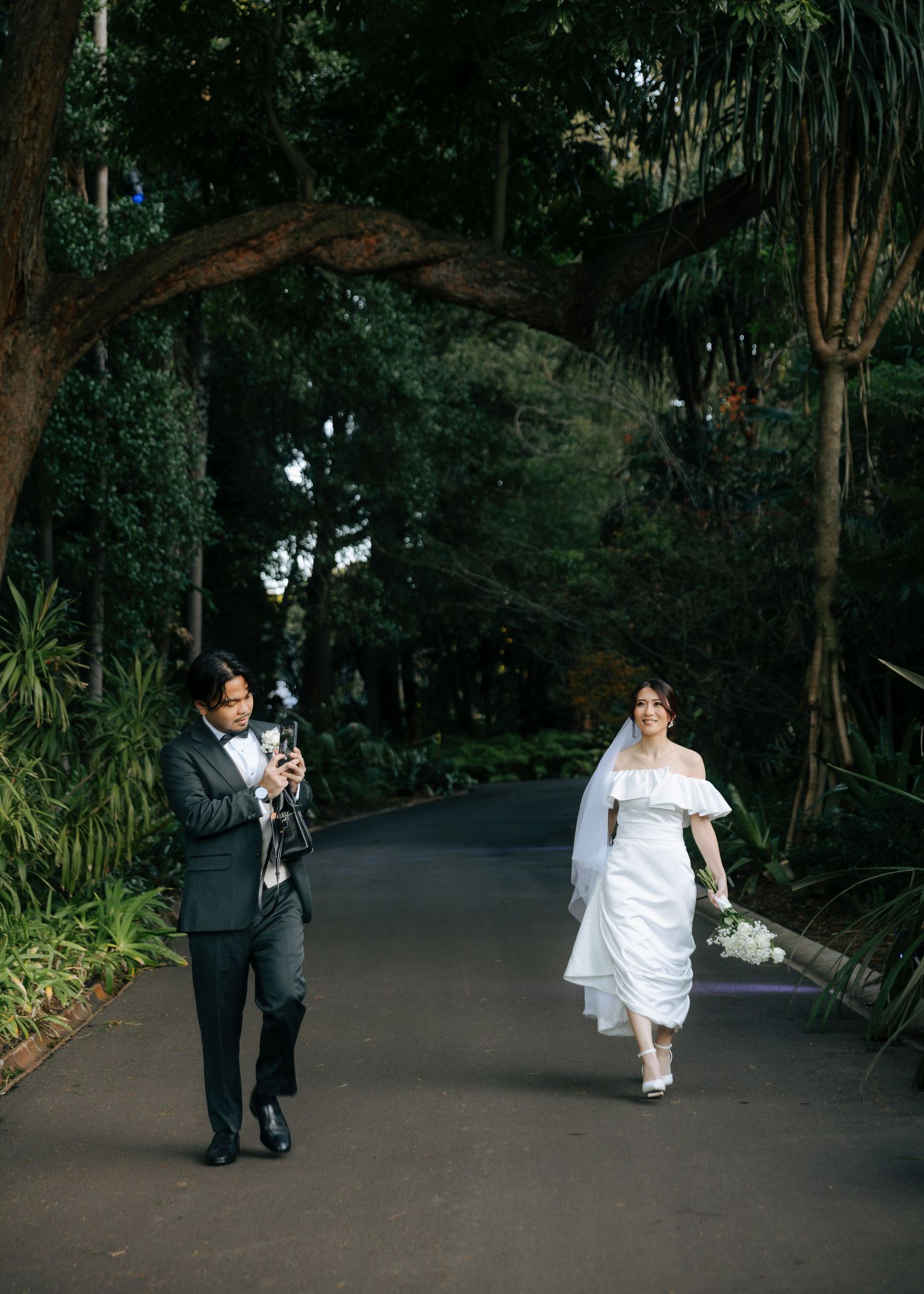 Bride in white off-shoulder gown and groom in dark suit walk along a tree-lined garden path.