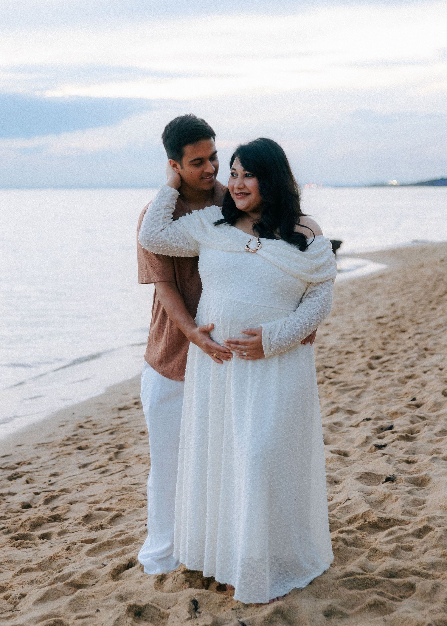 Couple maternity shoot on sandy beach at dusk, woman in white lace dress cradling baby bump.