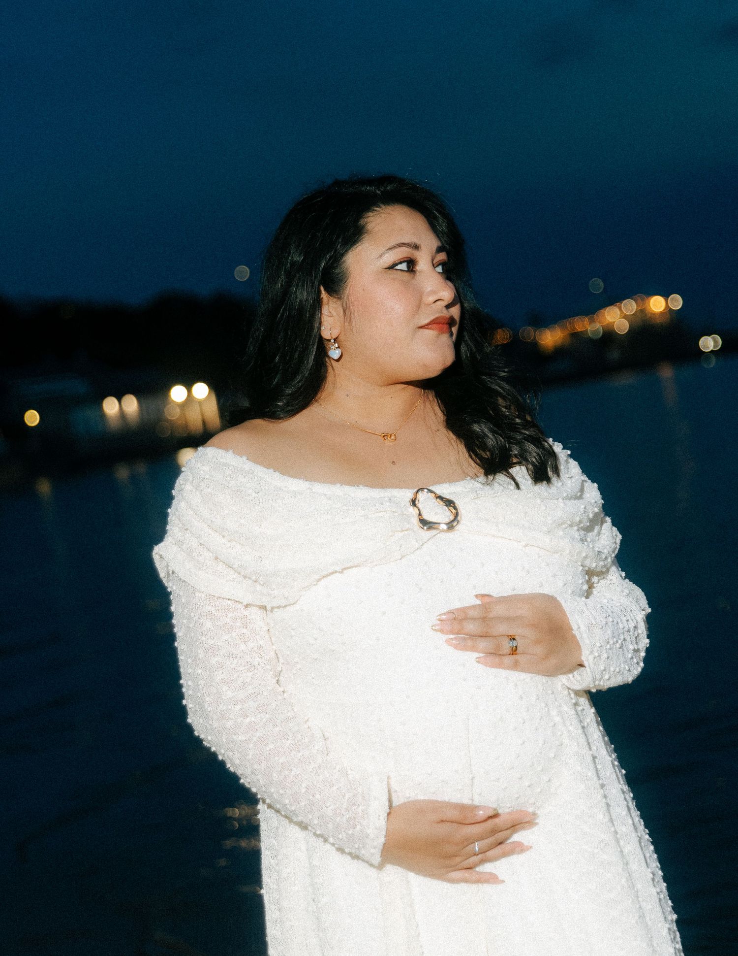 Pregnant woman in white textured dress holding belly near water with city lights at night.