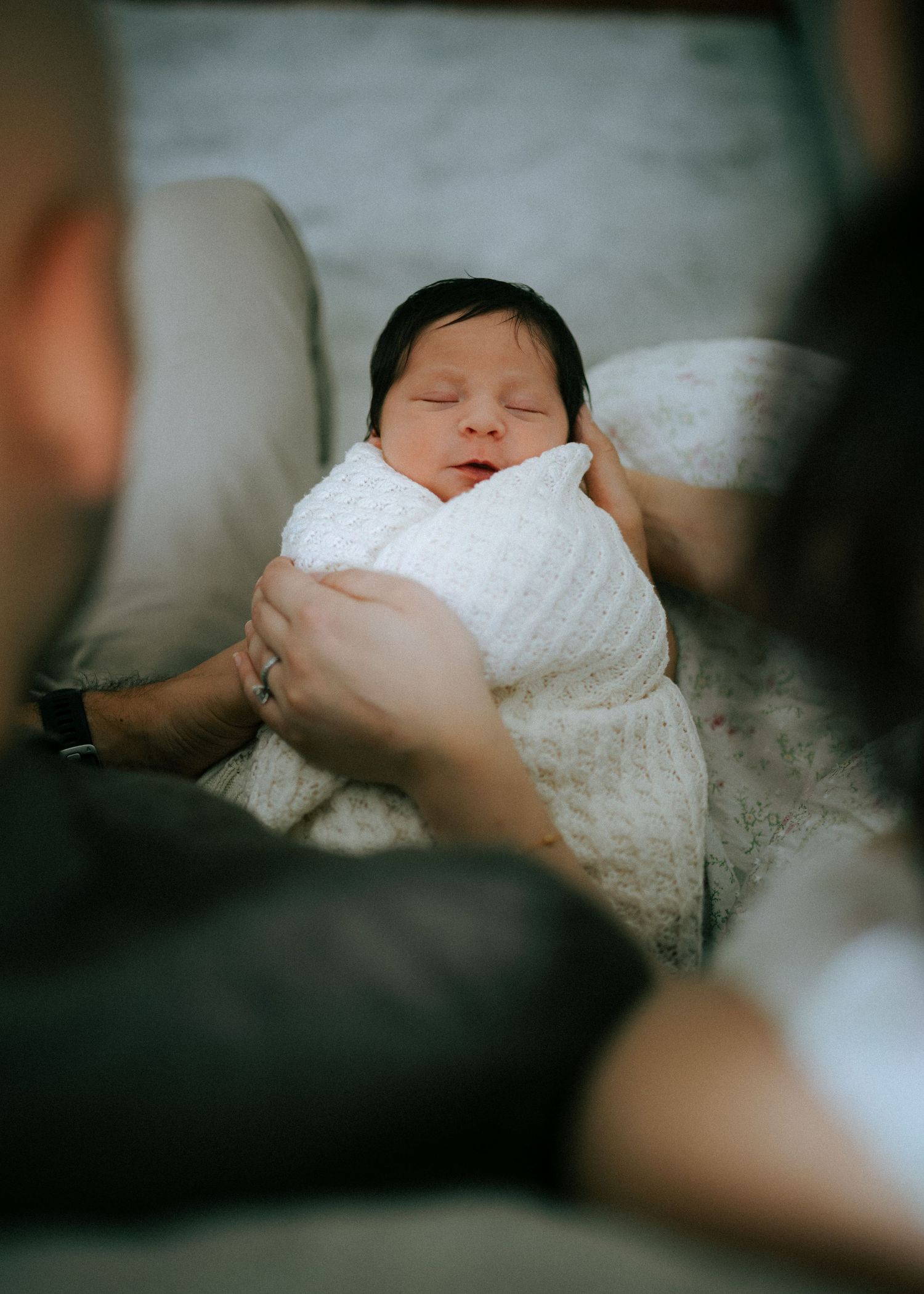 Newborn baby swaddled in white blanket sleeping peacefully while parents hold them gently.