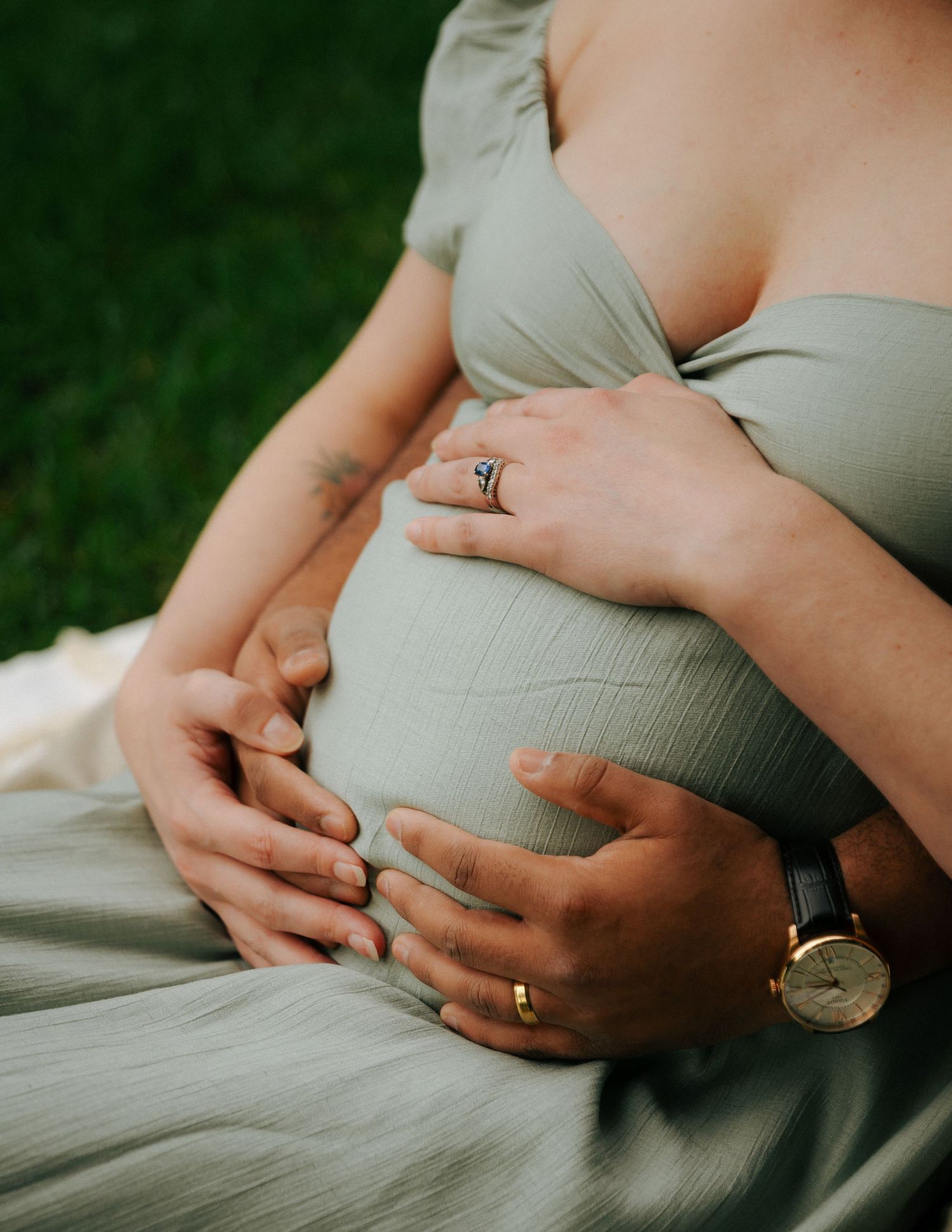 Close-up of couple's hands cradling pregnant belly, woman wearing engagement ring and green dress.