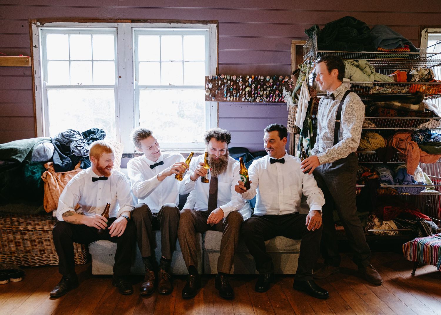 Groomsmen in white shirts and bow ties laughing and drinking beer together in a rustic wooden room before the wedding.