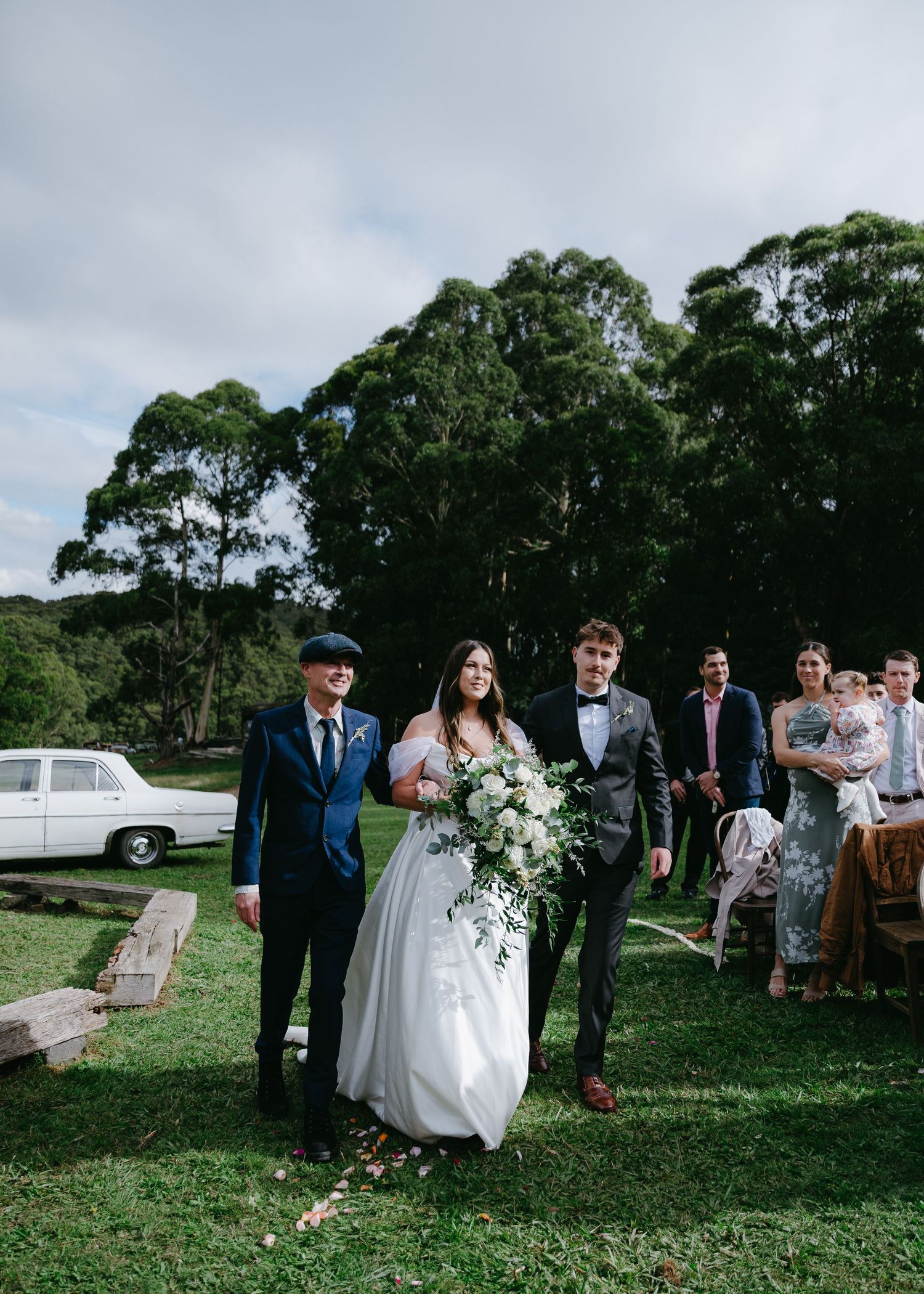 Bride walking down the aisle with two escorts holding a white bouquet at an outdoor wedding.