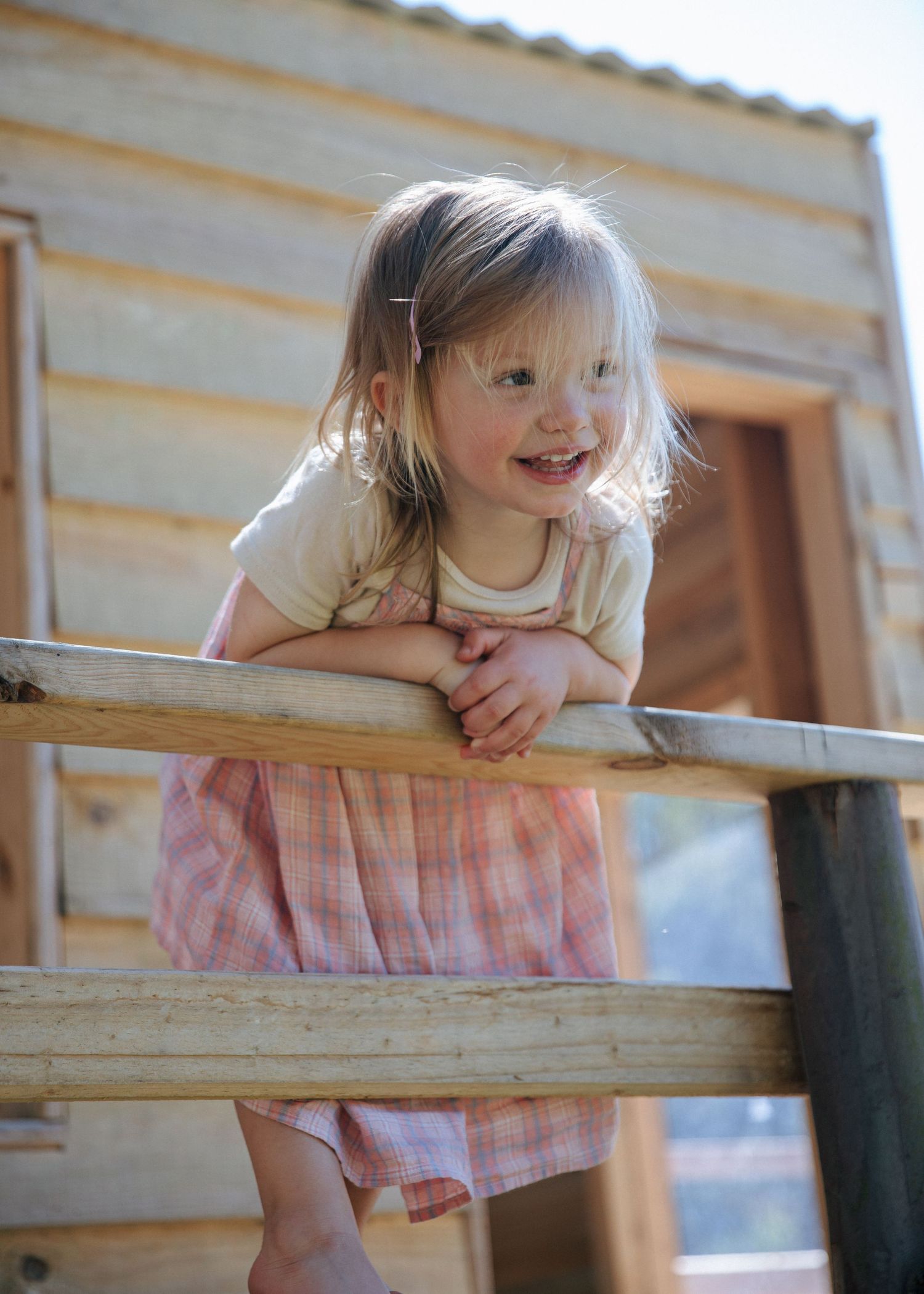 A smiling toddler girl in a pink dress leans on a wooden fence at a rustic playground.