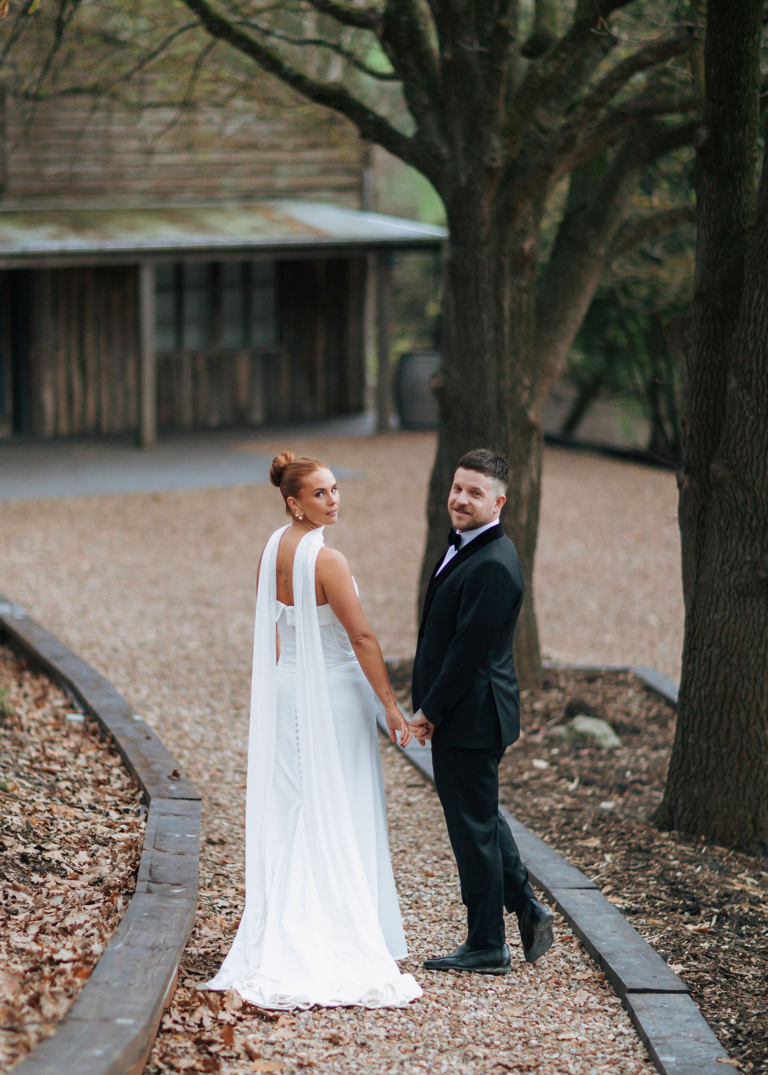 Bride and groom hold hands on a tree-lined path, looking back at the camera in elegant wedding attire.