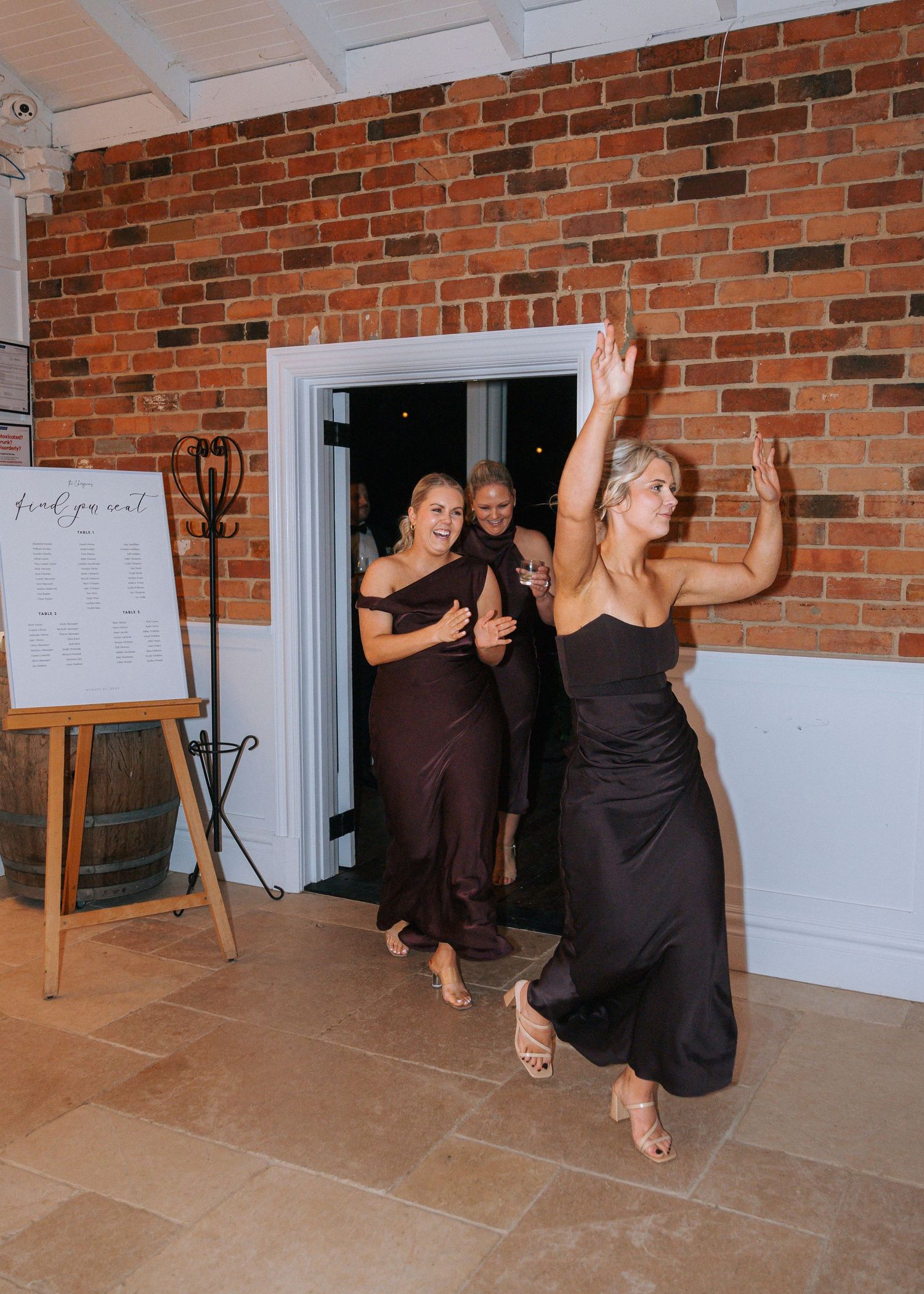 Three bridesmaids in dark satin gowns make celebratory entrance at wedding reception venue.
